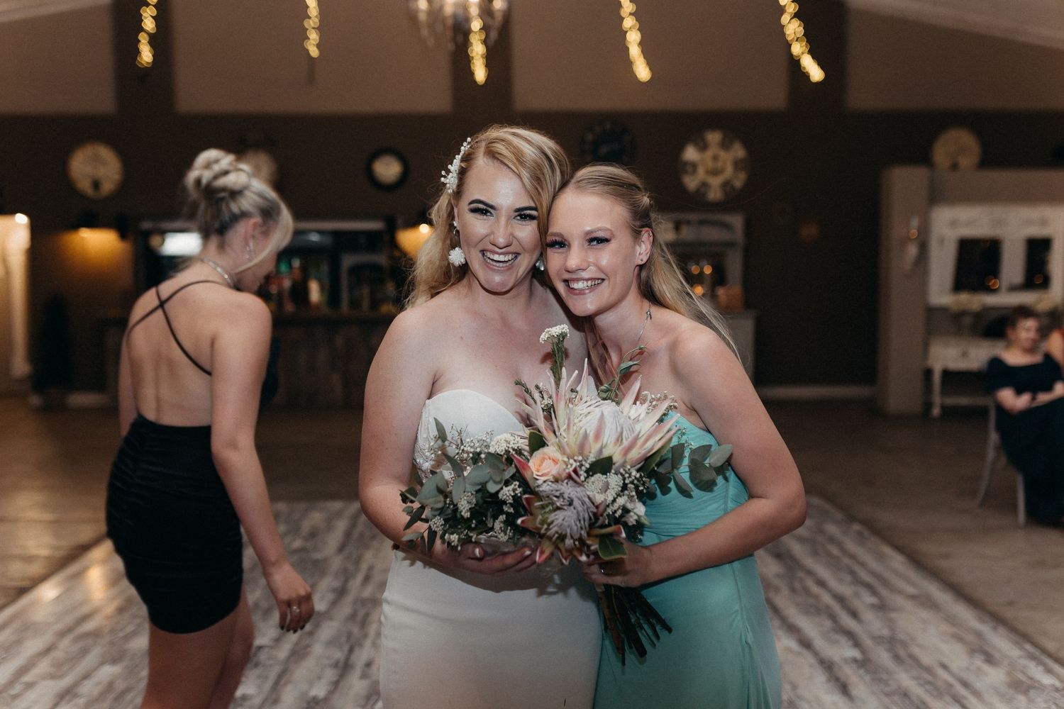 Two women embrace and smile while holding a floral bouquet at a wedding reception.
