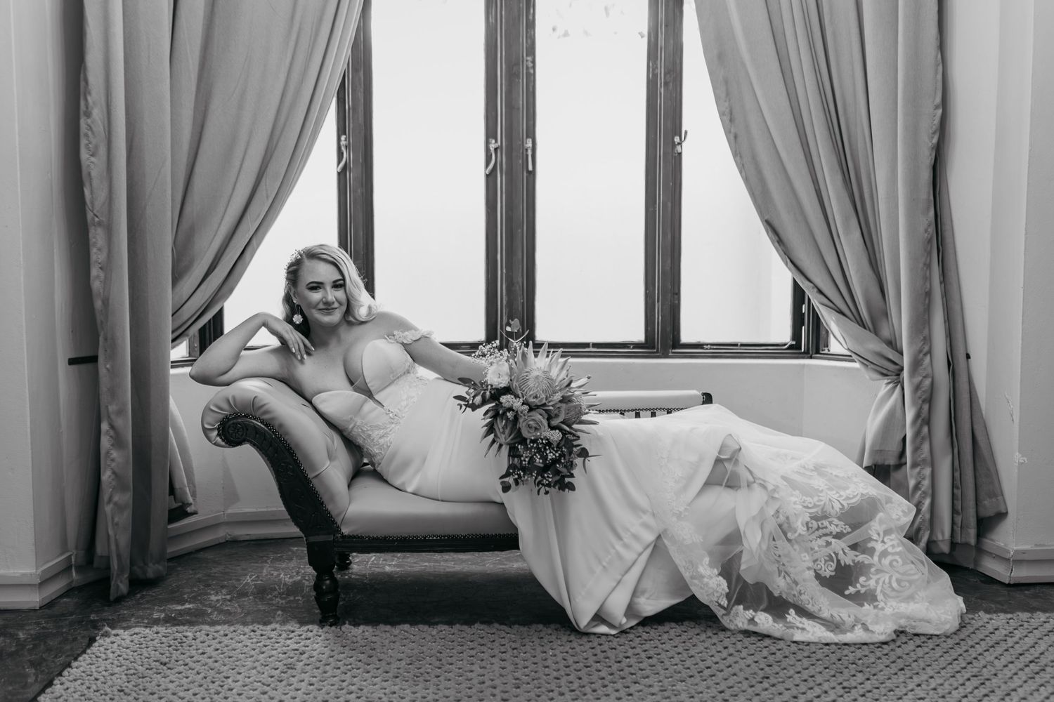 Black and white photo of bride reclining on vintage chaise lounge by curtained windows.