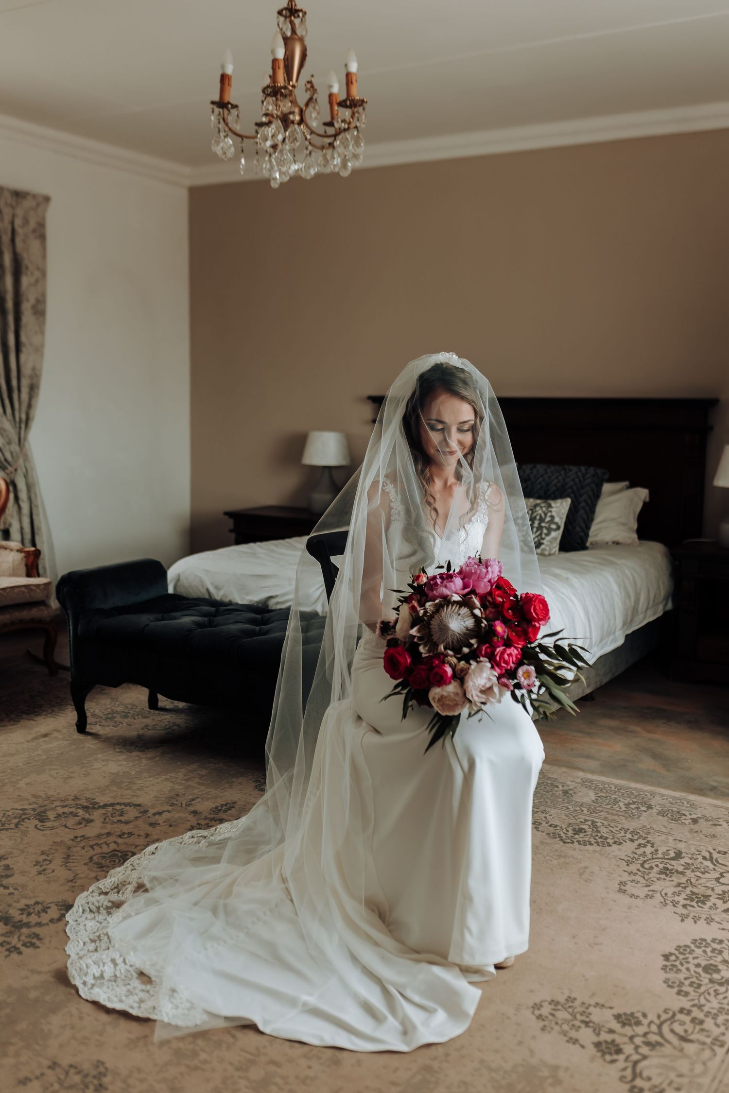 Bride in long veil sits with bouquet in elegant bedroom with chandelier.