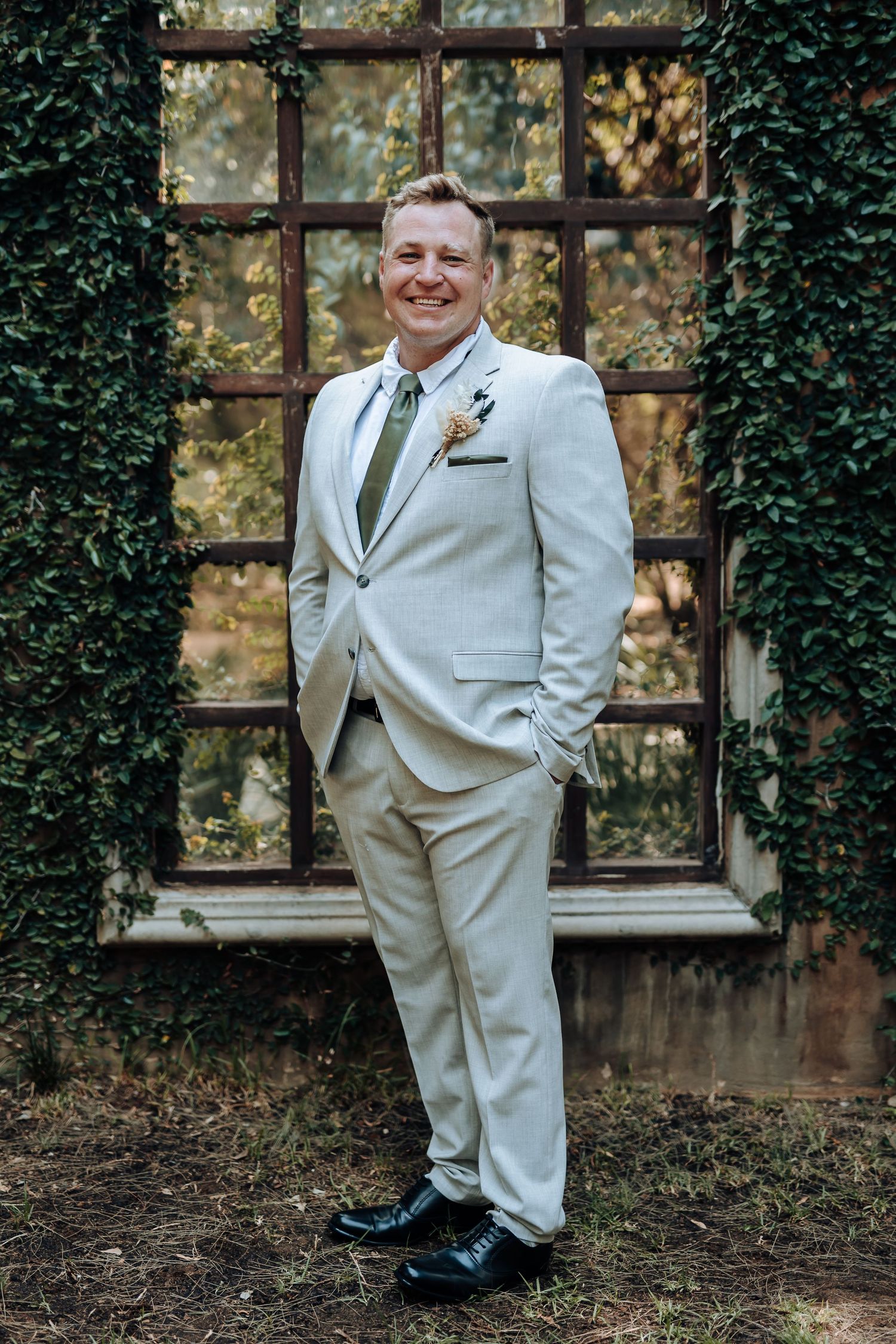 Person in a light suit poses confidently against an ivy-covered wall with large window frame.