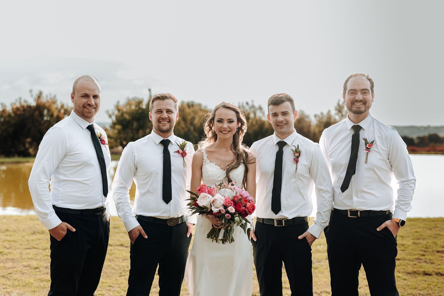 Wedding party in white shirts and black ties poses by lakeside setting.