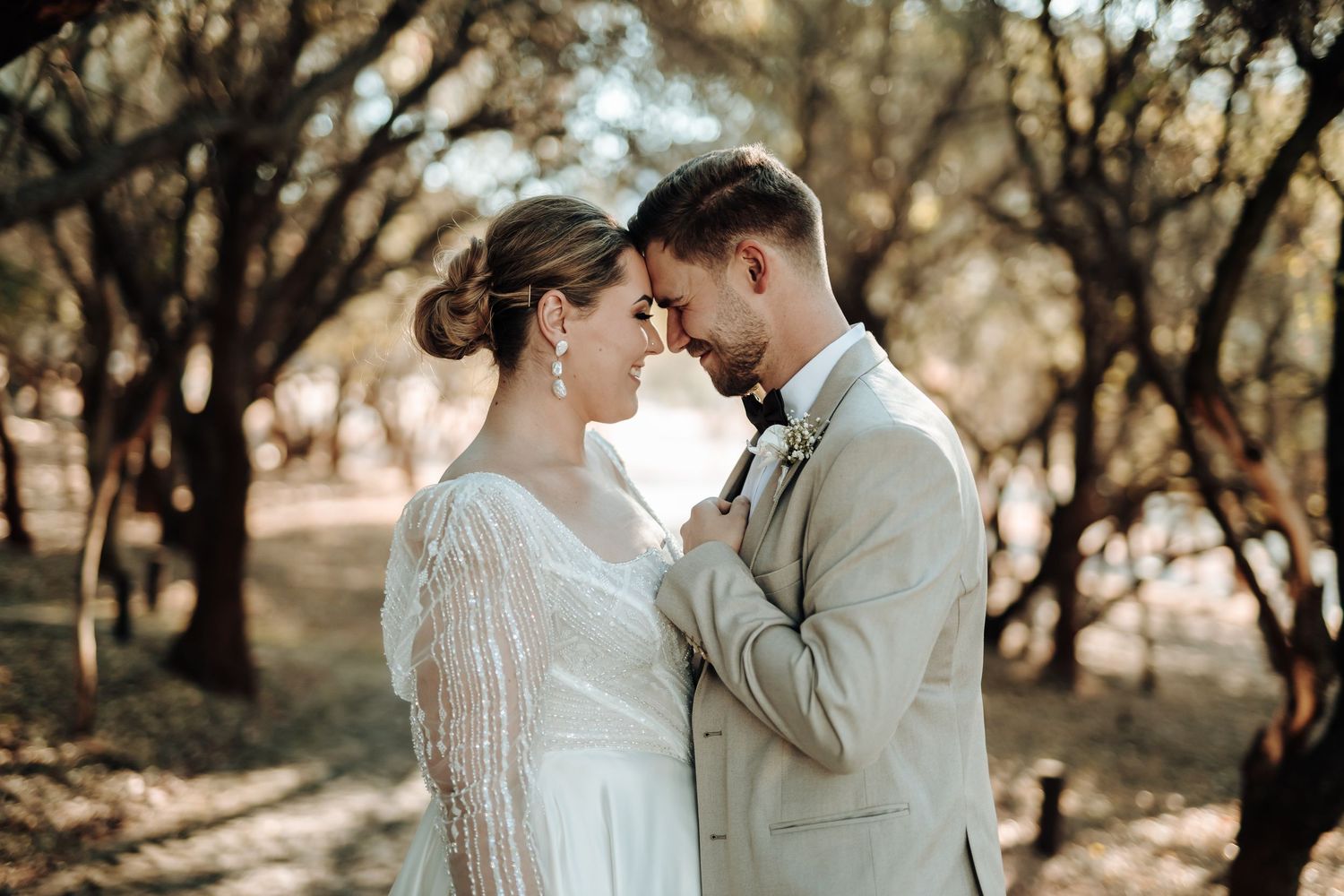 Tender moment between couple in elegant wedding attire under forest canopy.