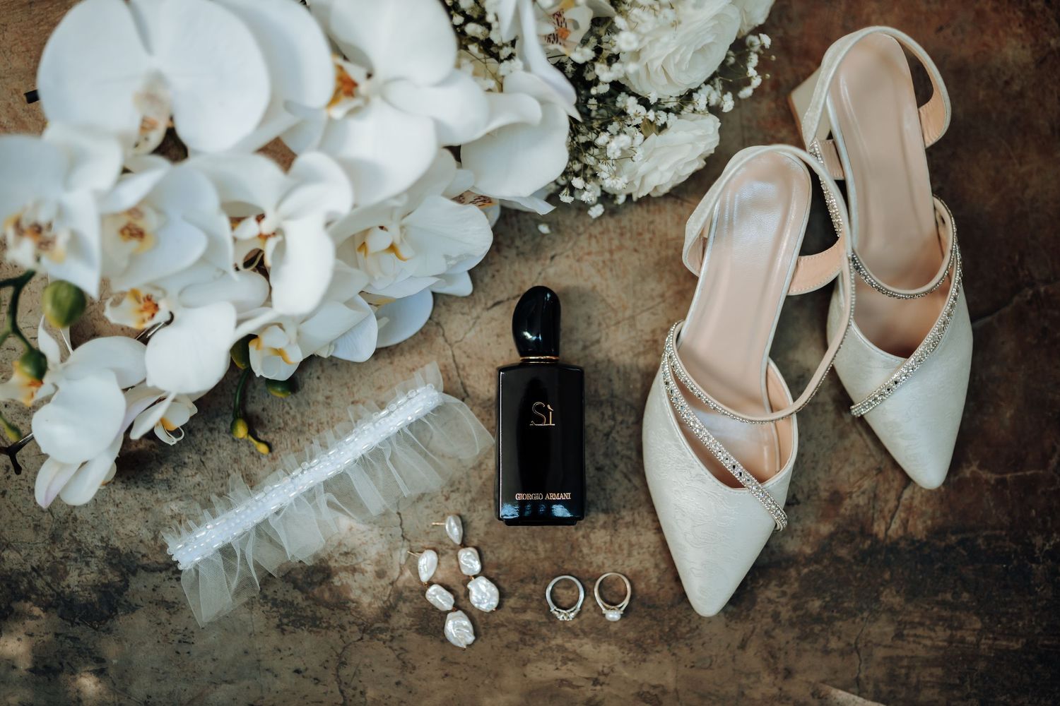 Wedding detail flatlay with white heels, orchids, perfume bottle, and jewelry on wooden surface.