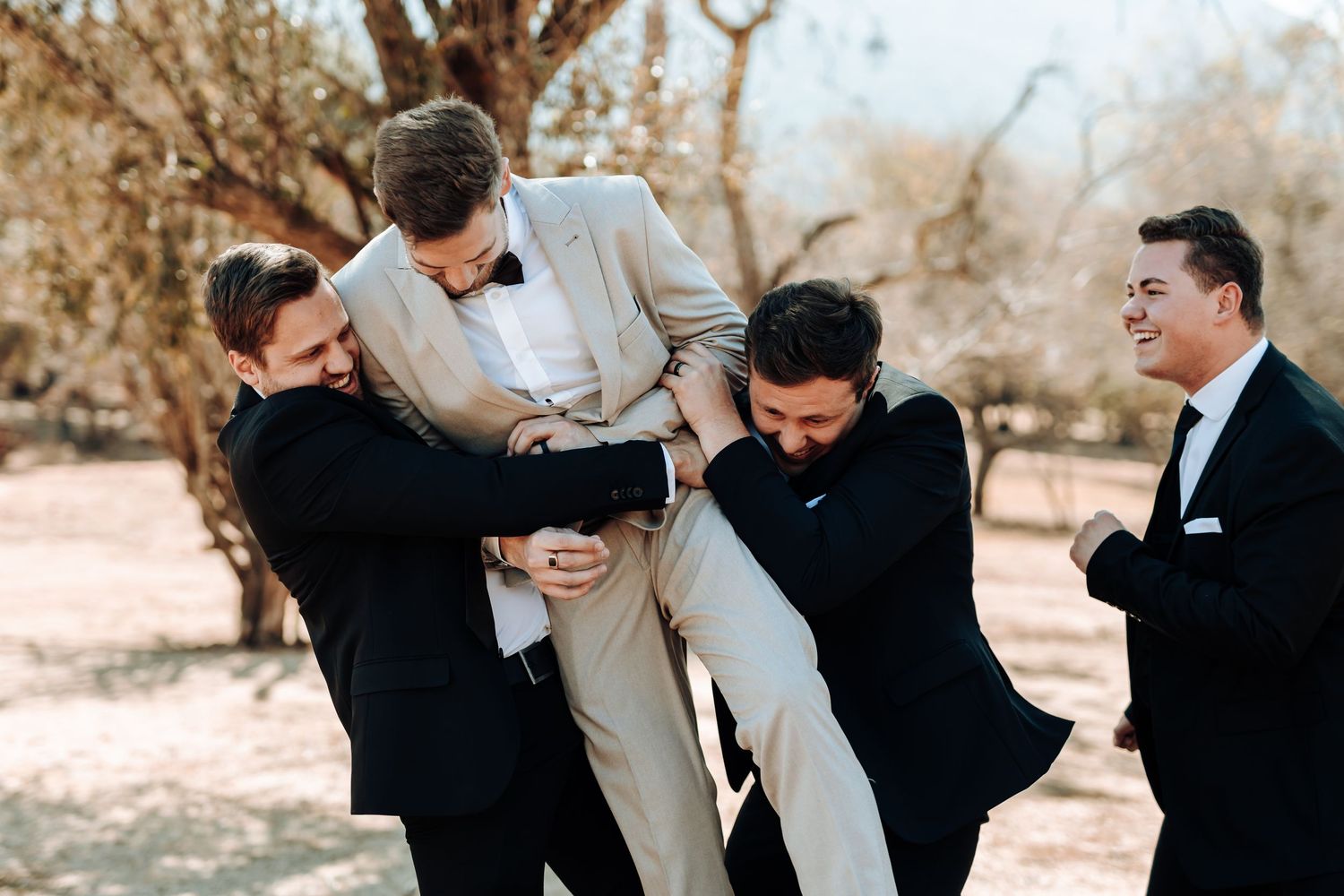 Groomsmen in black suits playfully lift their friend in a light colored suit during a wedding photoshoot.