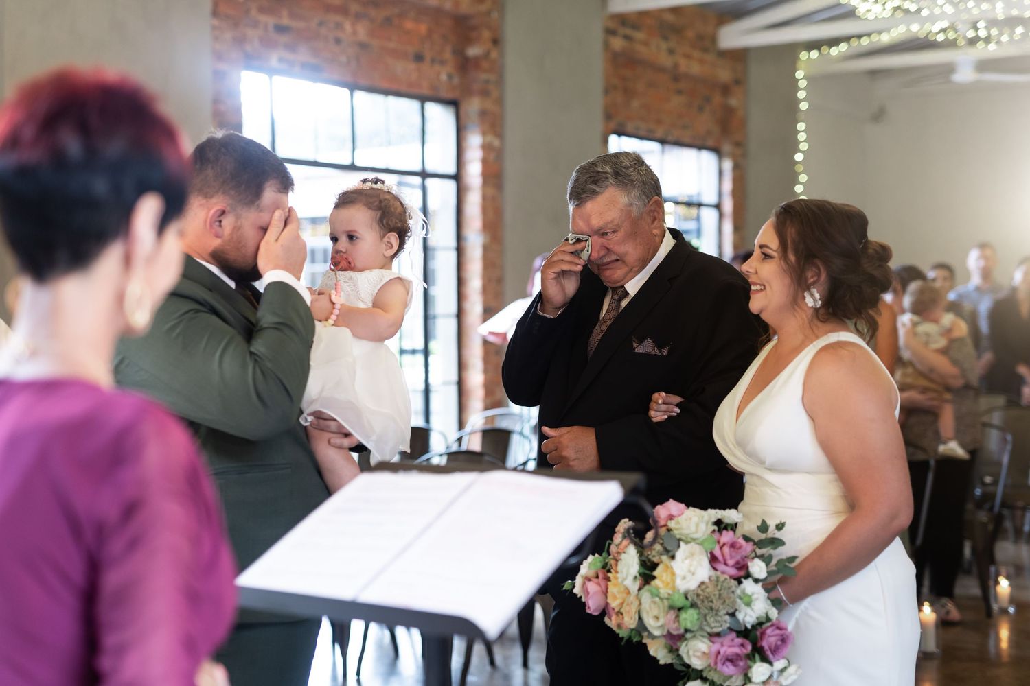 An indoor wedding ceremony scene with exposed brick walls and a small baby present.
