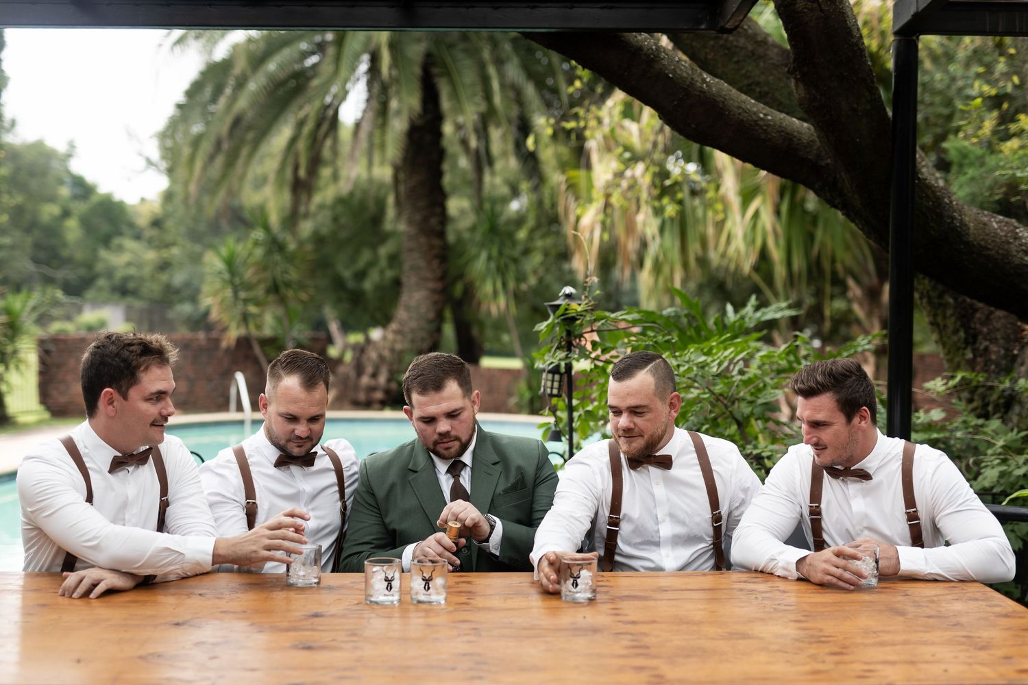 Five wedding party members share drinks at a wooden table in a tropical garden setting.