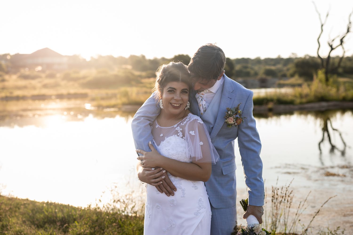 Wedding couple embraces during golden hour by lake with natural landscape.