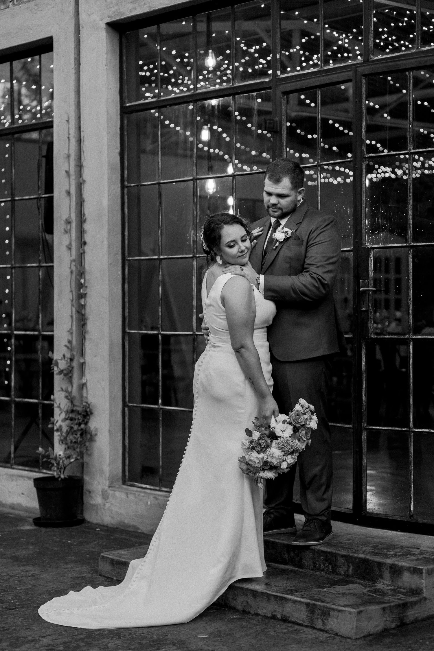Black and white portrait of couple in formal attire against window backdrop with string lights.
