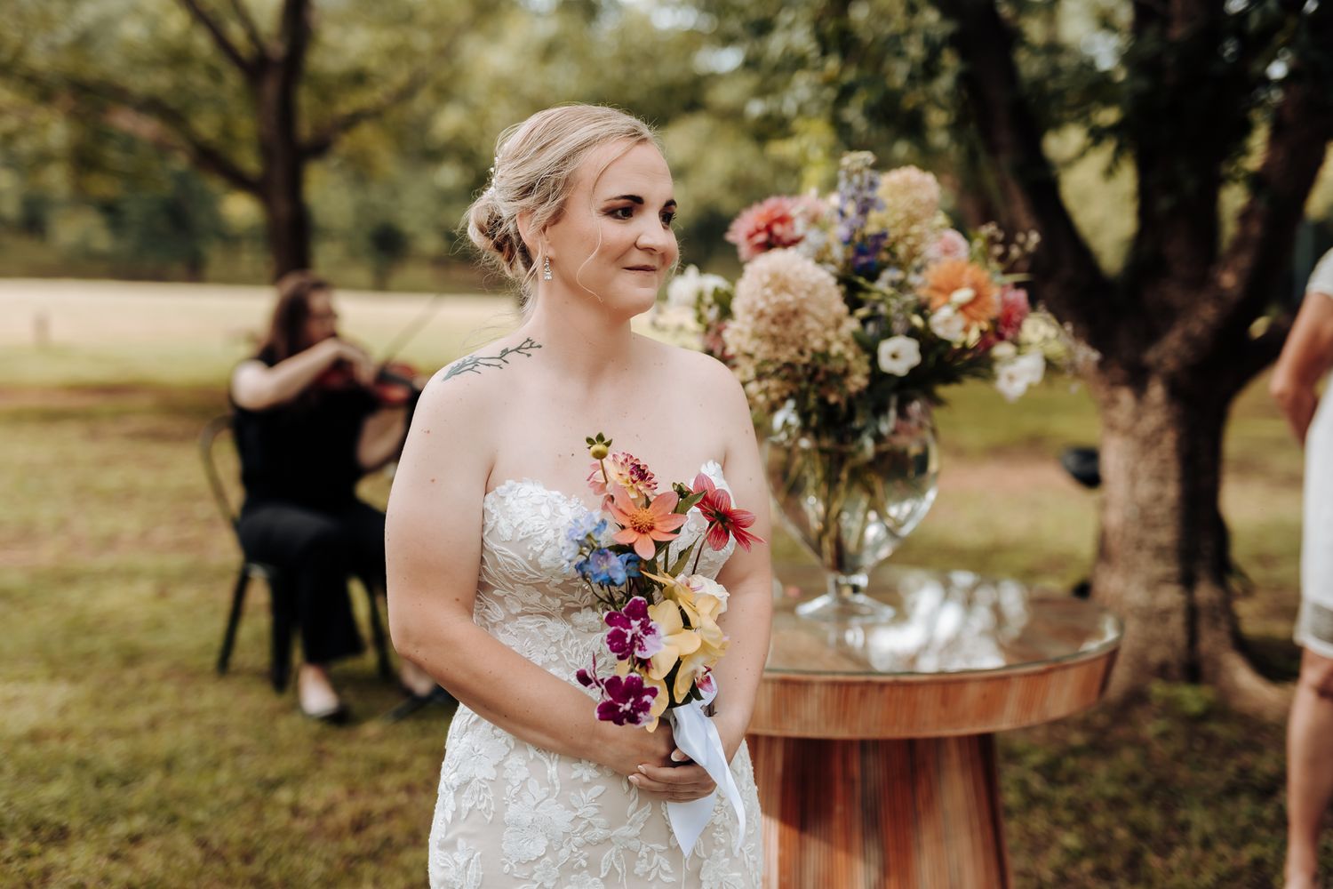Person in lace wedding dress holds colorful bouquet while standing in outdoor garden setting.