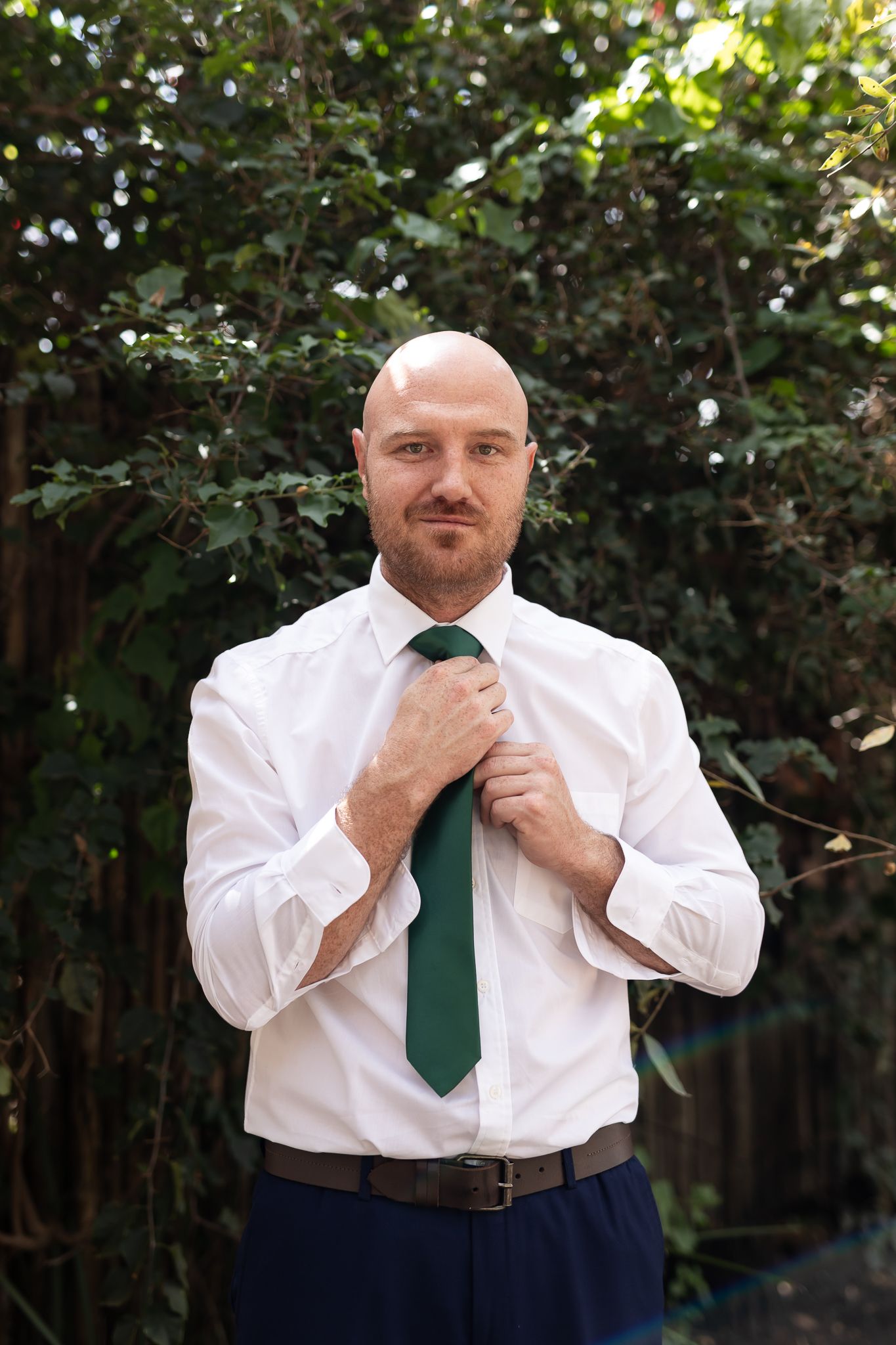Man adjusting green tie while wearing white dress shirt and navy pants outdoors.