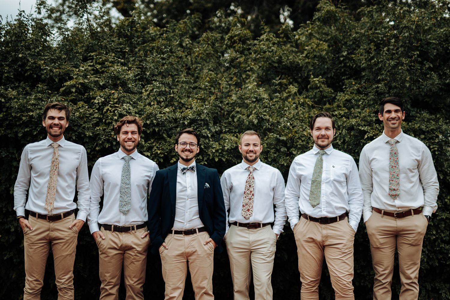 Groomsmen and groom in coordinated attire pose together against green hedge backdrop.