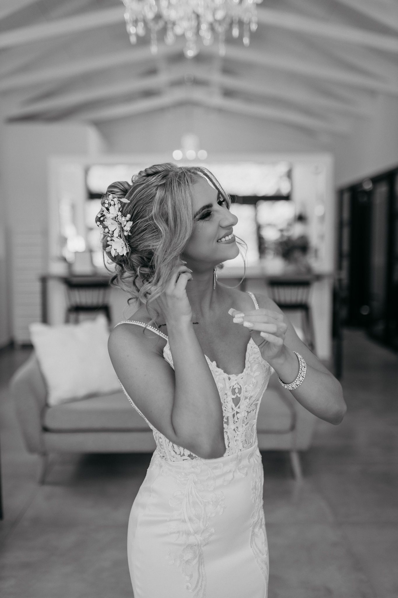 Black and white photo of person in wedding dress adjusting jewelry beneath elegant chandelier.