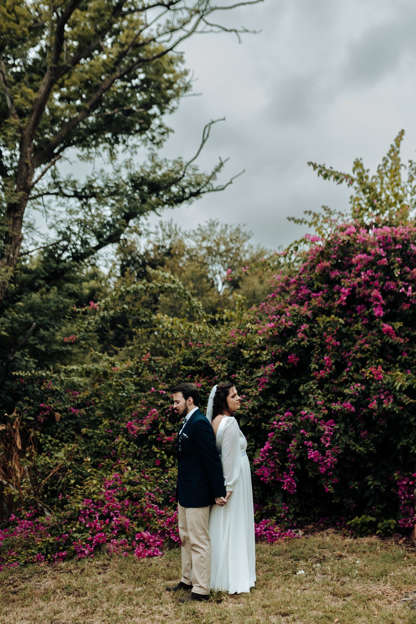 Wedding couple poses against dramatic sky and pink flowering bushes in garden setting.
