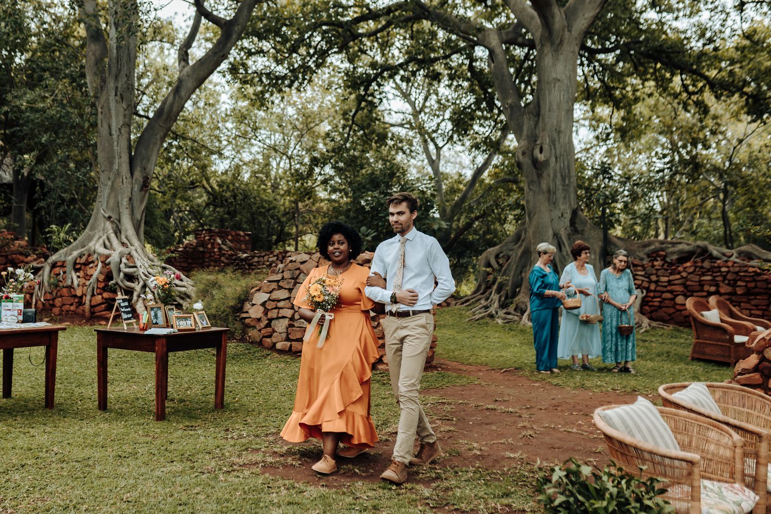 Guests in colorful attire take part in an outdoor wedding processional on a grassy lawn.