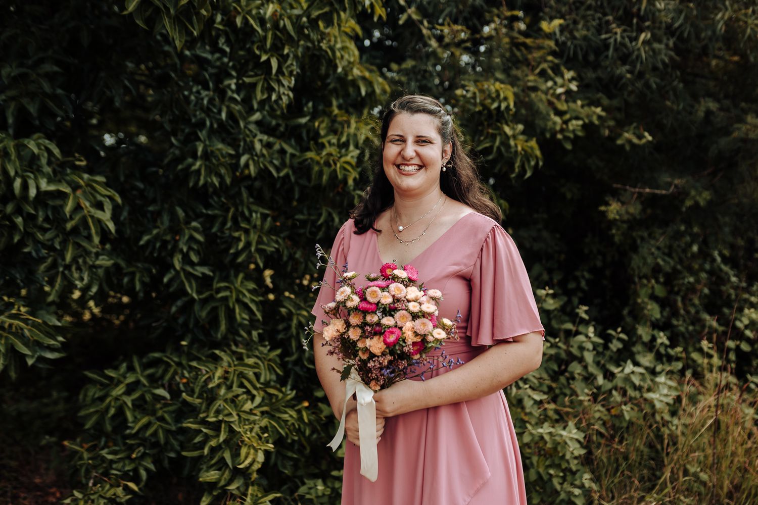 Person in dusty rose dress holding pink floral bouquet in outdoor garden setting.