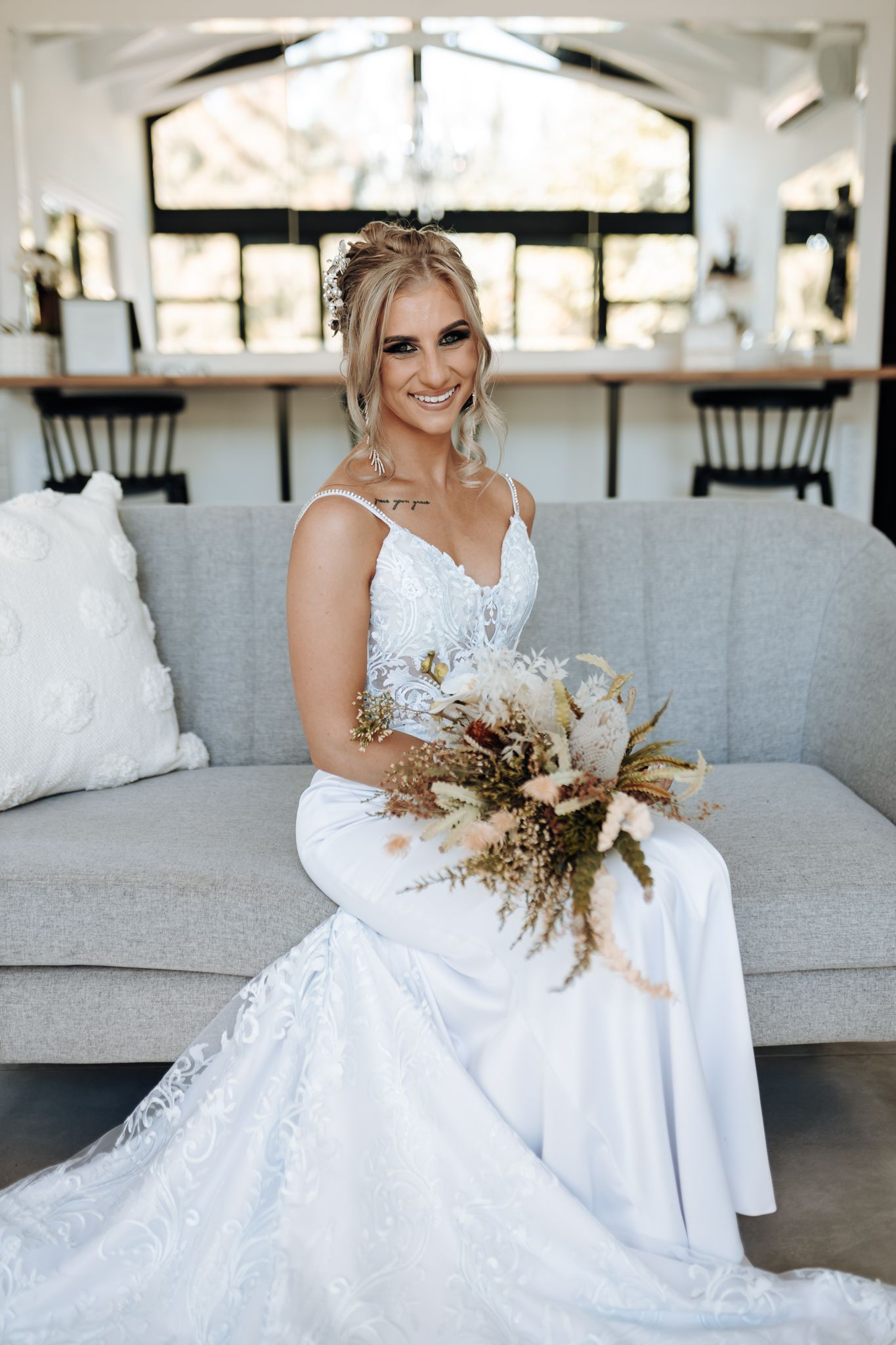 Bride holding dried floral bouquet while sitting on gray sofa under crystal chandelier.