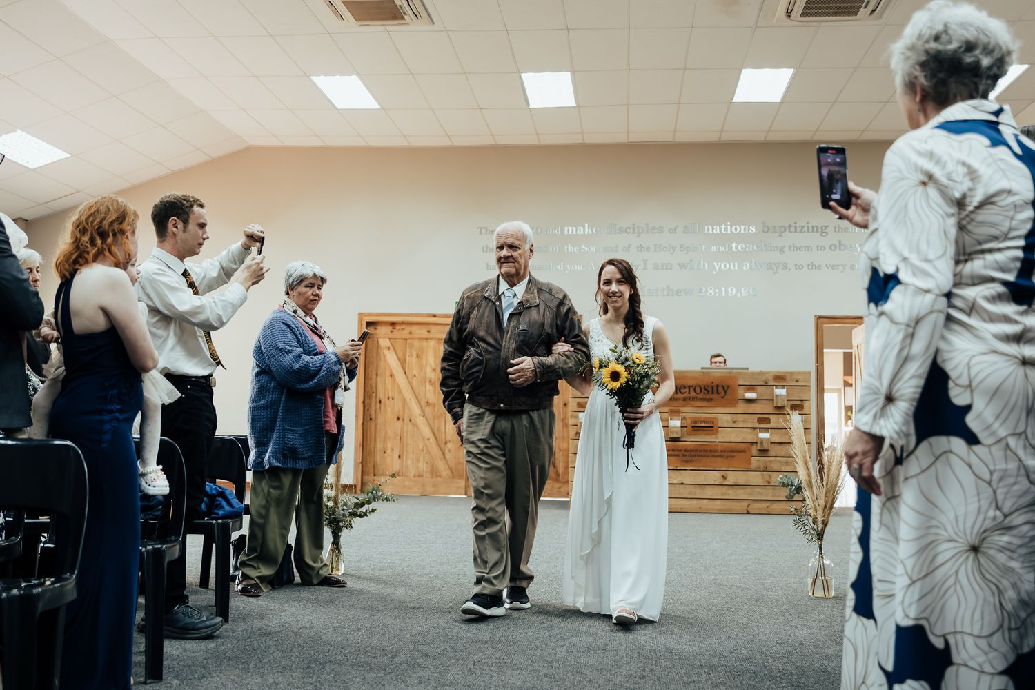 Wedding guests gather in a bright indoor reception venue with fluorescent lighting and wooden accents.