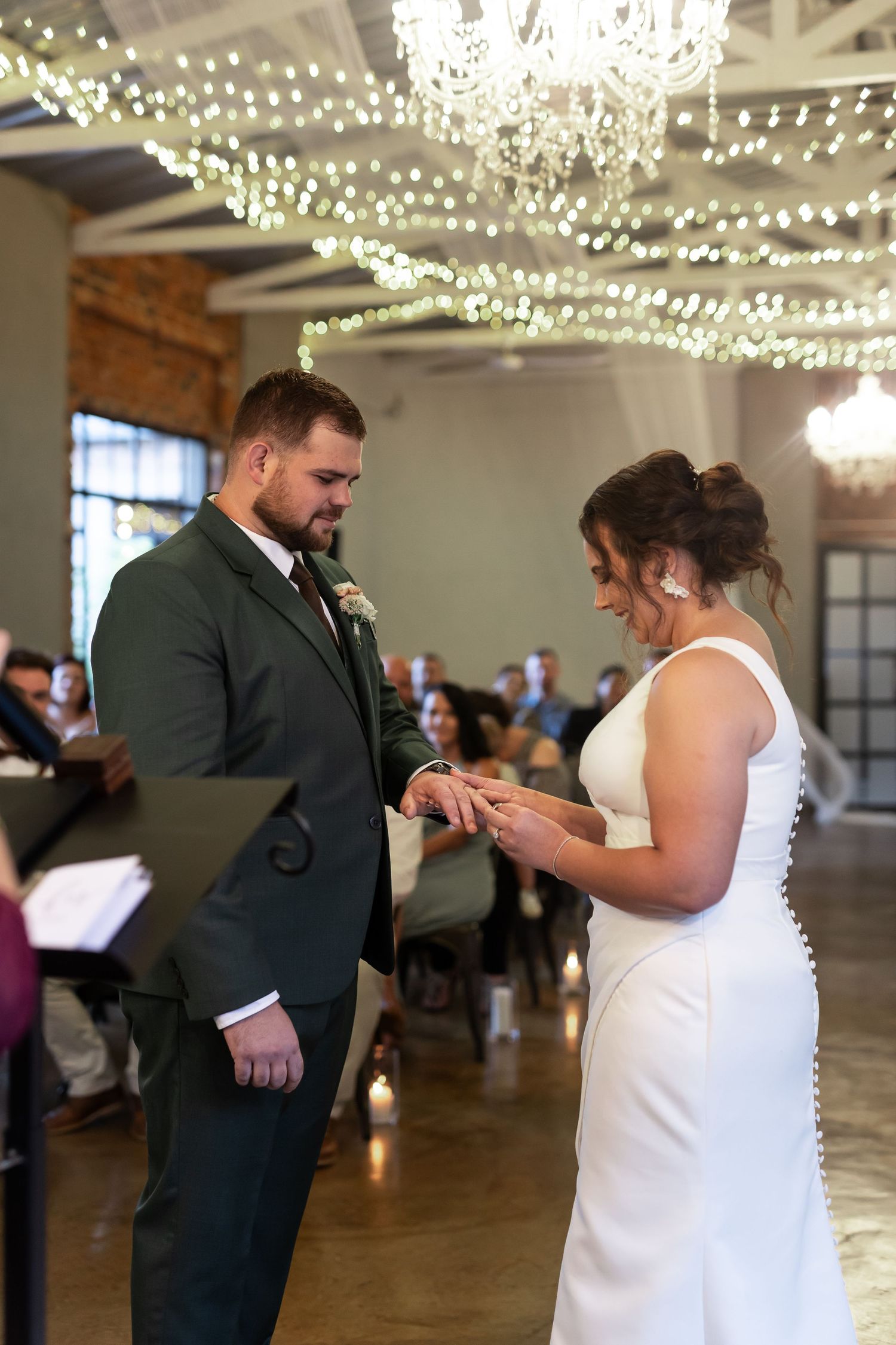 Ring exchange during wedding ceremony under crystal chandeliers and twinkle lights.
