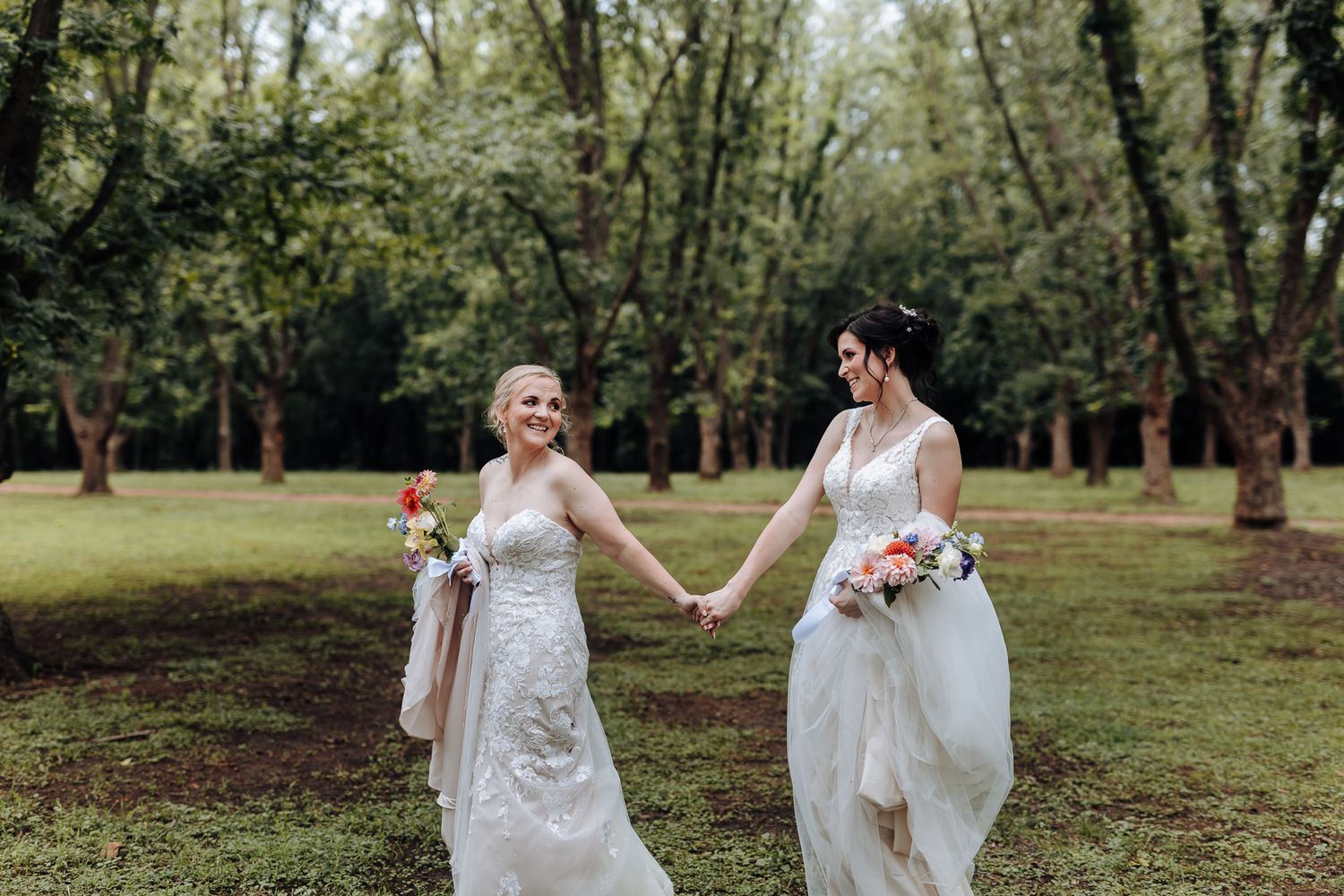 Two brides in white lace dresses hold hands while walking through a scenic wooded park.