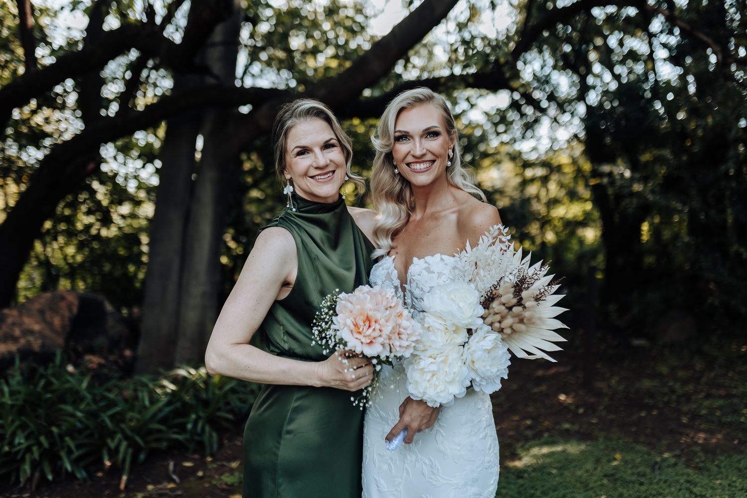 Wedding portrait of two elegantly dressed people with bouquets featuring white and blush flowers in a garden setting.