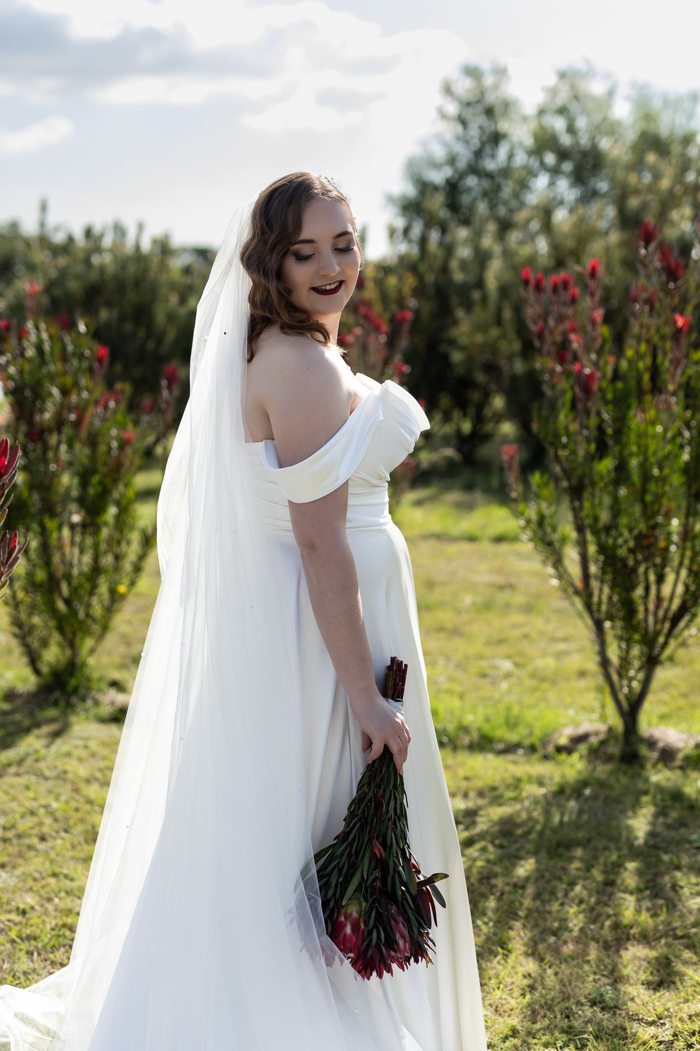 Bride in off-shoulder gown looks over shoulder while holding bouquet in garden setting.