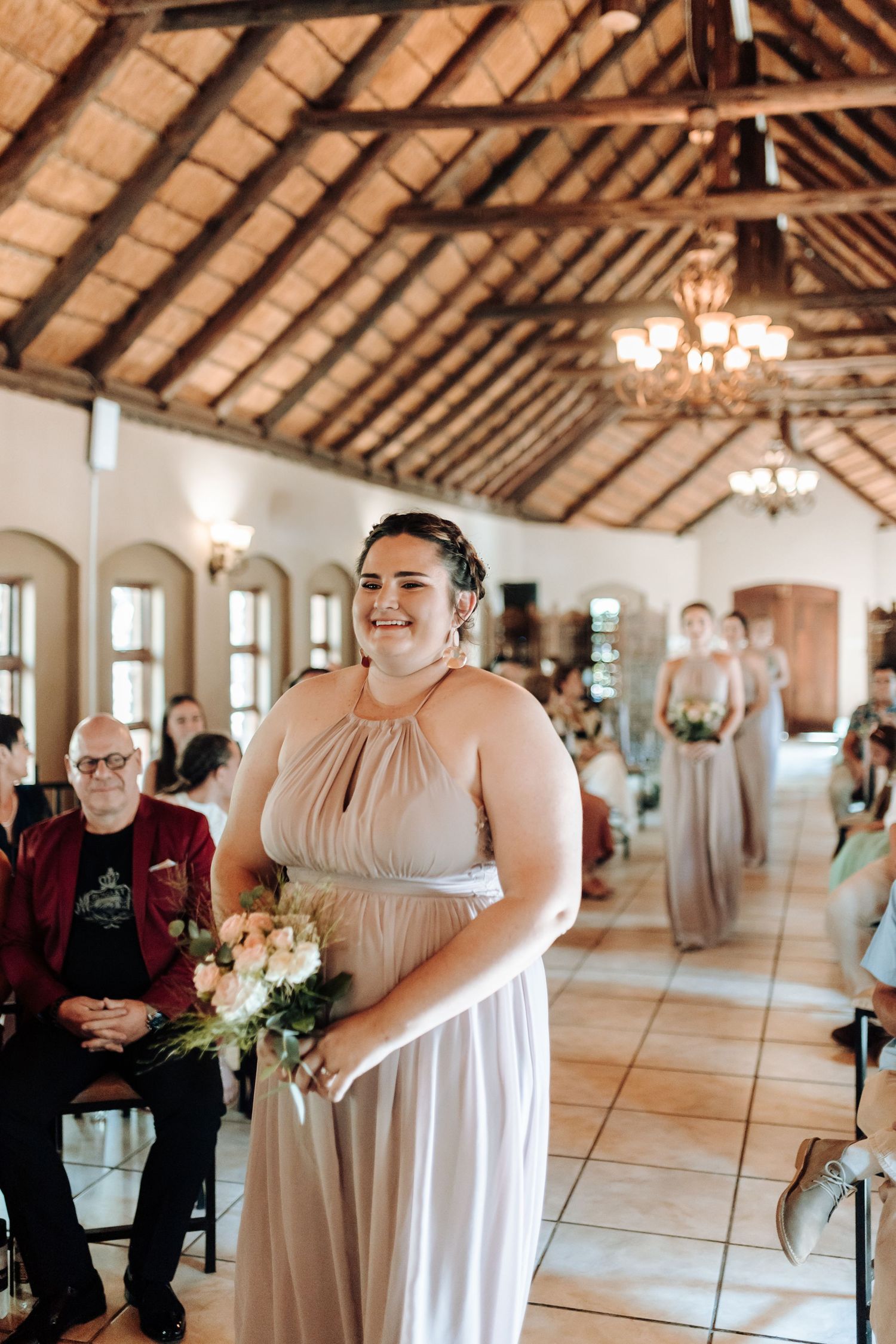 A bridesmaid in a champagne-colored halter dress stands holding a bouquet in a rustic venue with exposed beams.