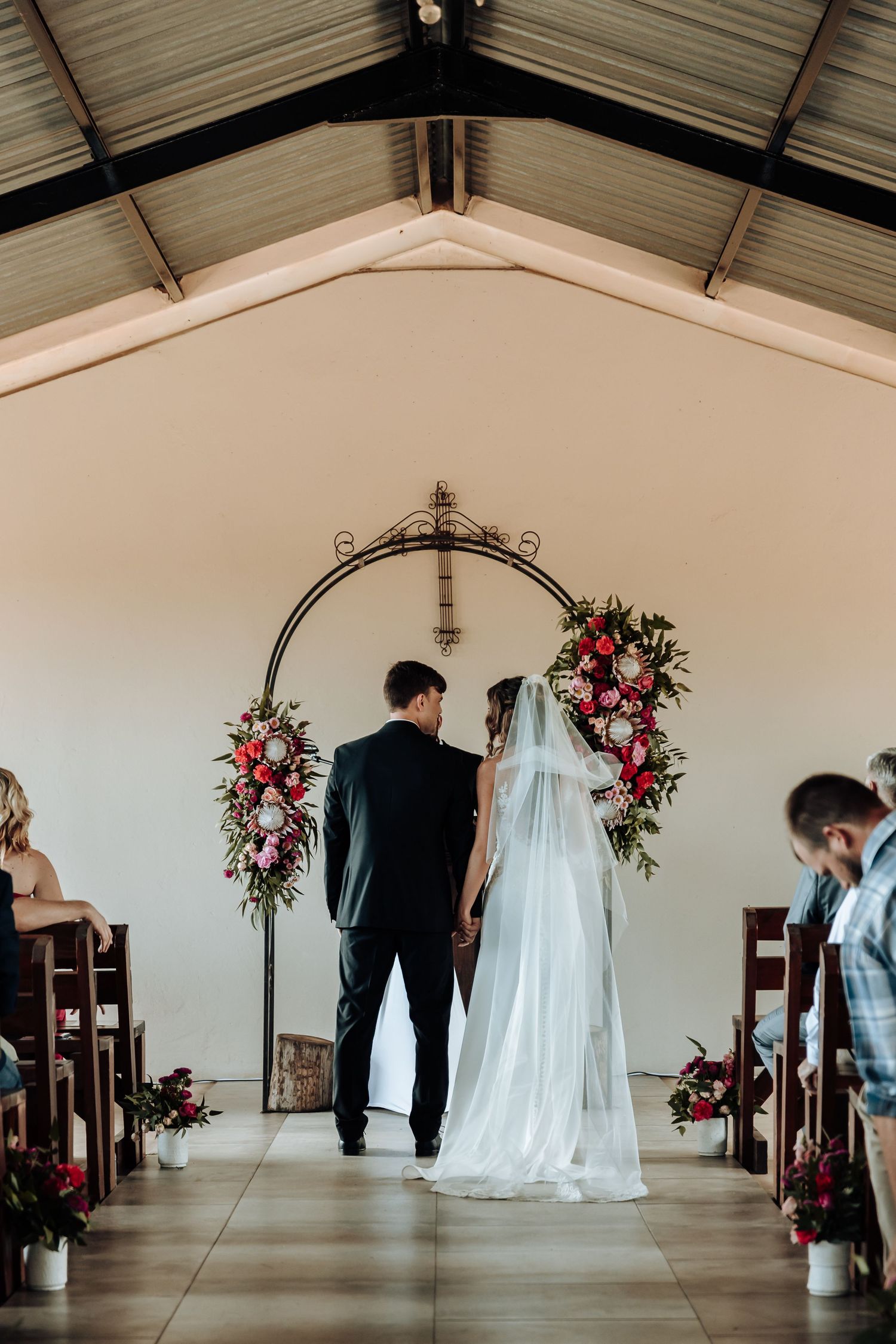 Wedding couple stands at altar beneath arched floral installation with pink roses.