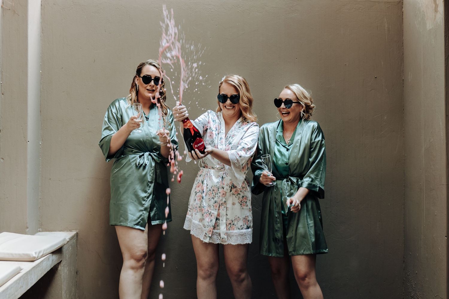 Friends in satin robes pop champagne and celebrate together against a neutral wall.