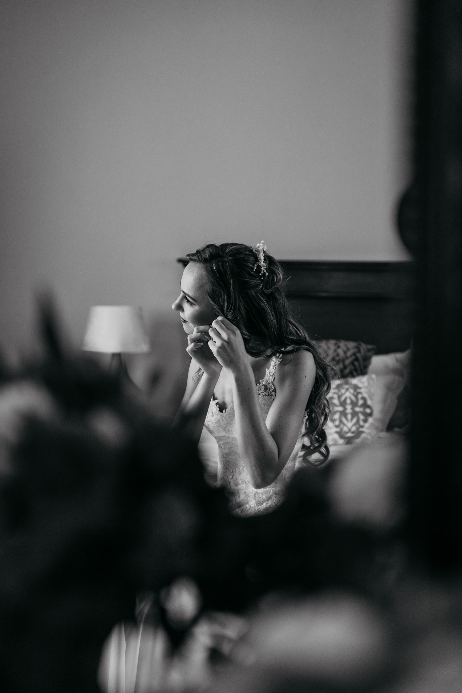Black and white photo of bride putting on earrings in elegant bedroom setting.