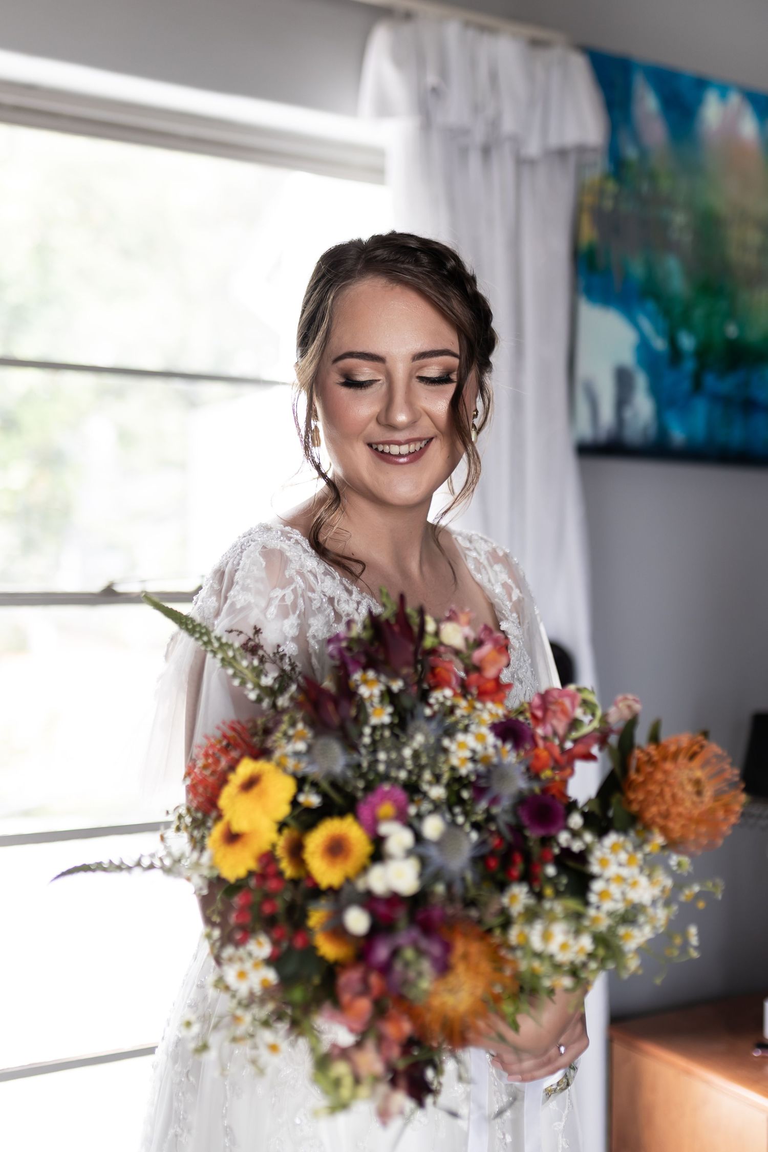 A bride in a lace wedding dress smiles while holding a vibrant autumn-colored bouquet by a window.
