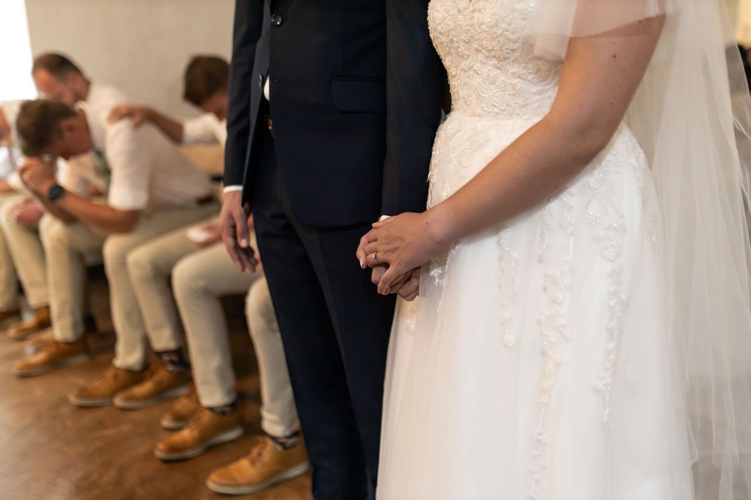 Close-up of formal wedding attire showing a black suit and white lace dress with groomsmen in background.