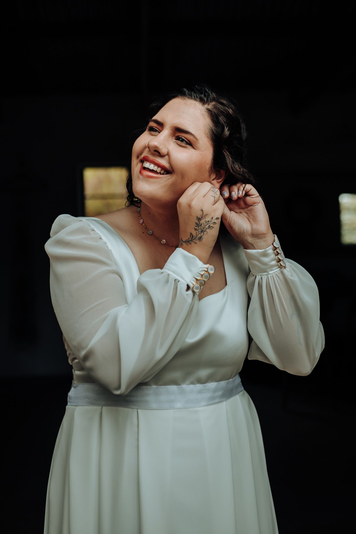 Portrait of a person in a white dress with sheer sleeves smiling while touching their earring.