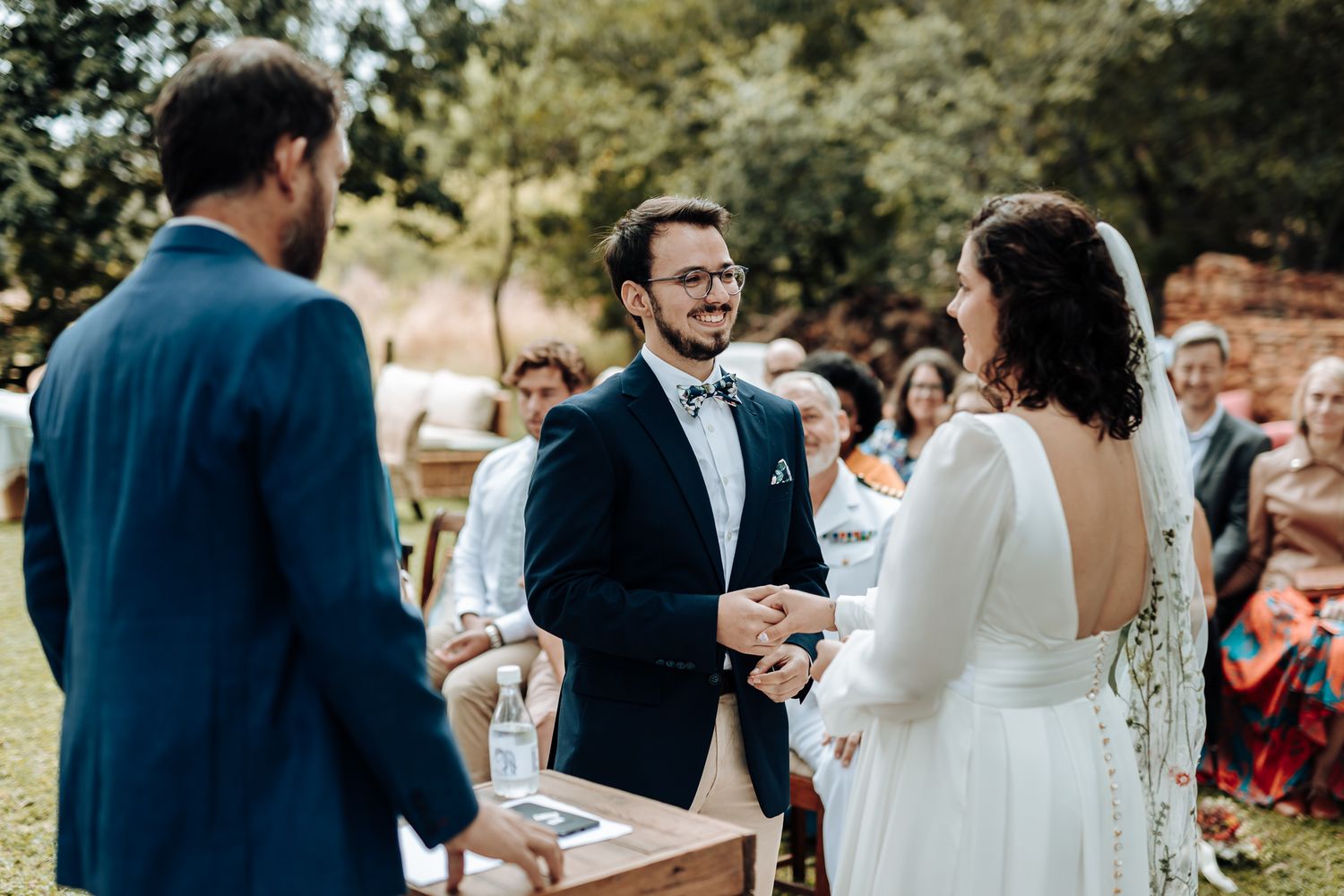 An outdoor wedding ceremony shows a couple exchanging vows in front of seated guests on a sunny day.