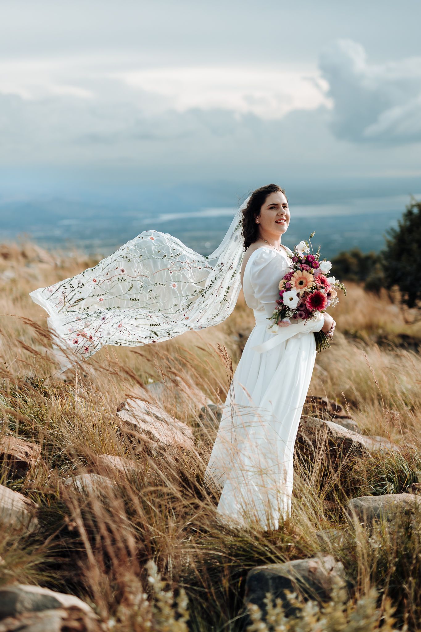 A bride in a flowing white lace dress holds a pink bouquet against a mountainous landscape.