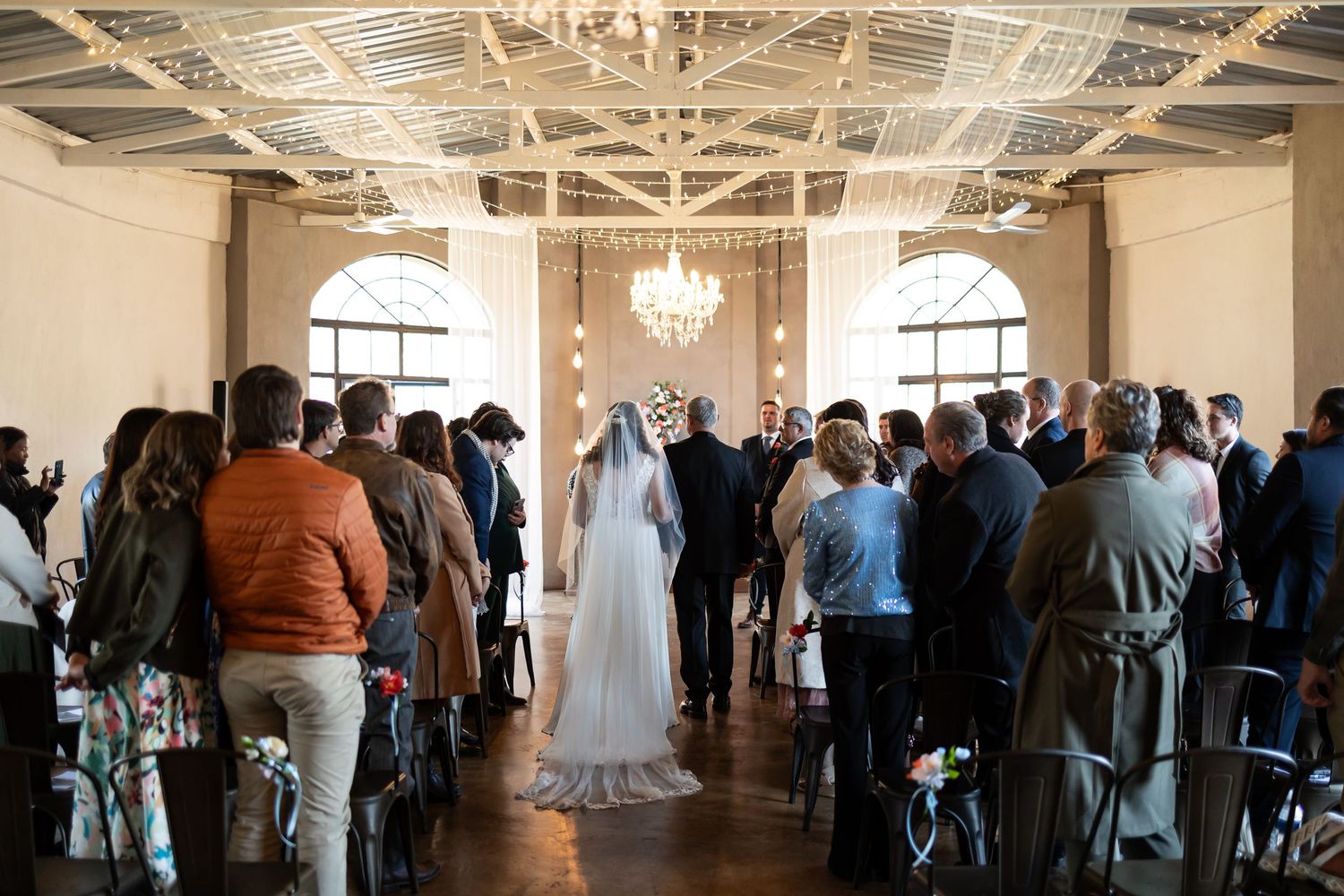 A wedding ceremony takes place in an elegant indoor venue with crystal chandeliers and exposed industrial ceiling details.