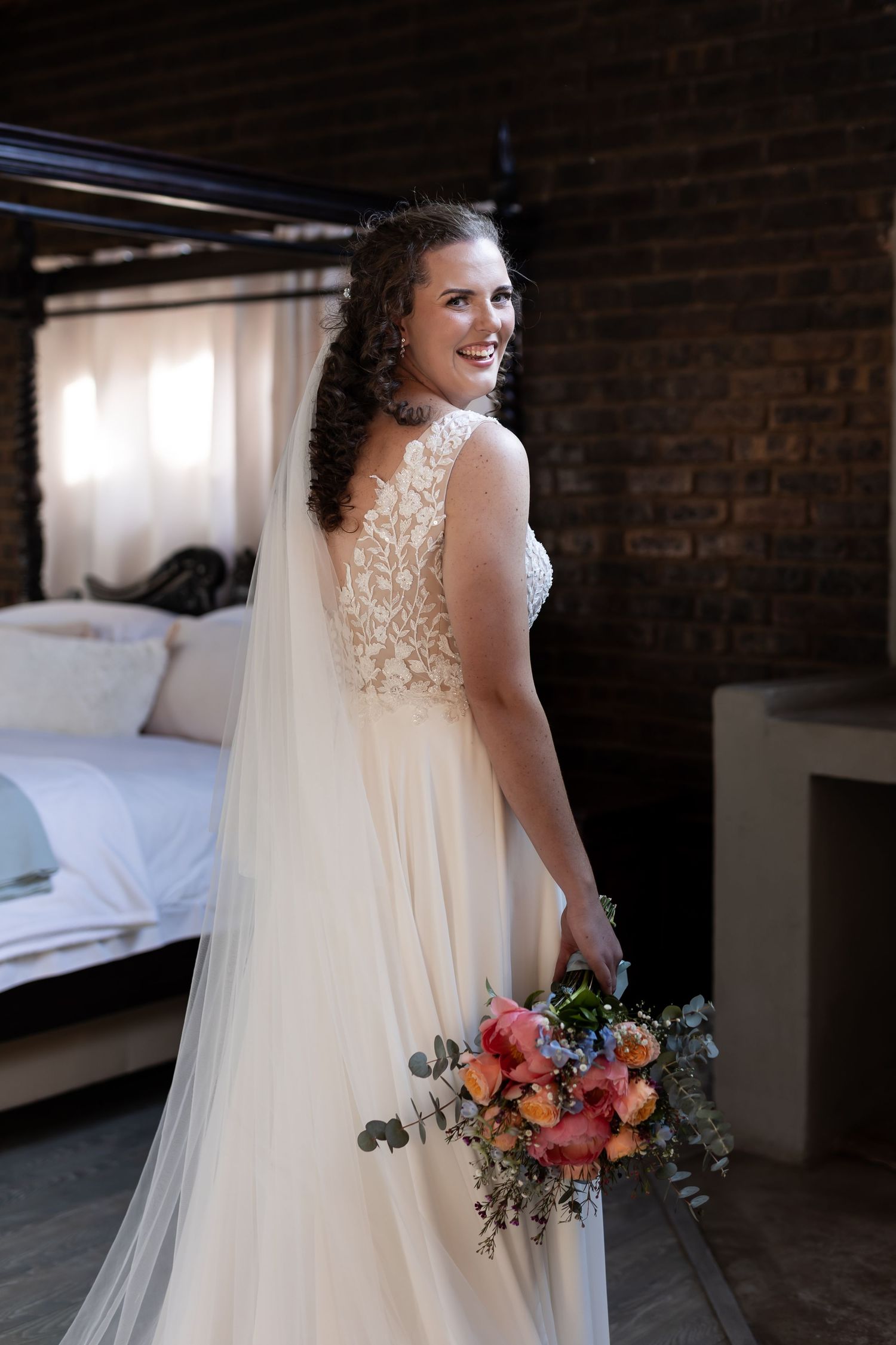 A bride in a flowing white lace dress holds a vibrant coral and blue floral bouquet against exposed brick walls.