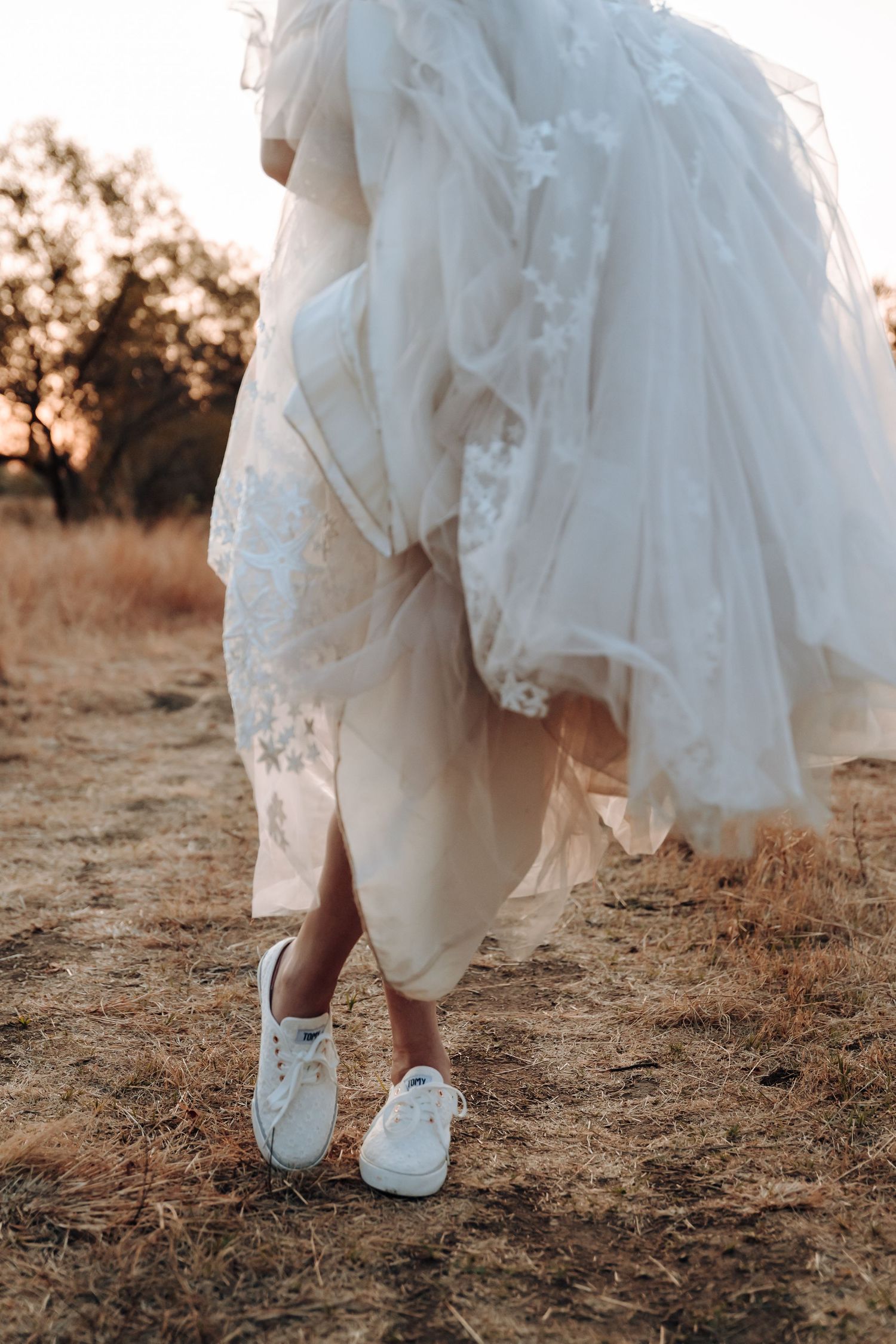 Bride wearing comfortable sneakers under her elegant lace wedding dress in outdoor setting.