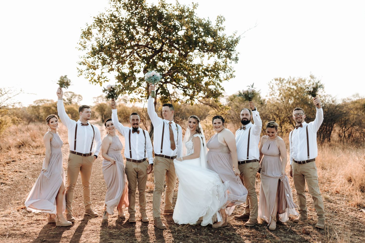 Joyful wedding party jumps and celebrates in outdoor setting with rustic backdrop.