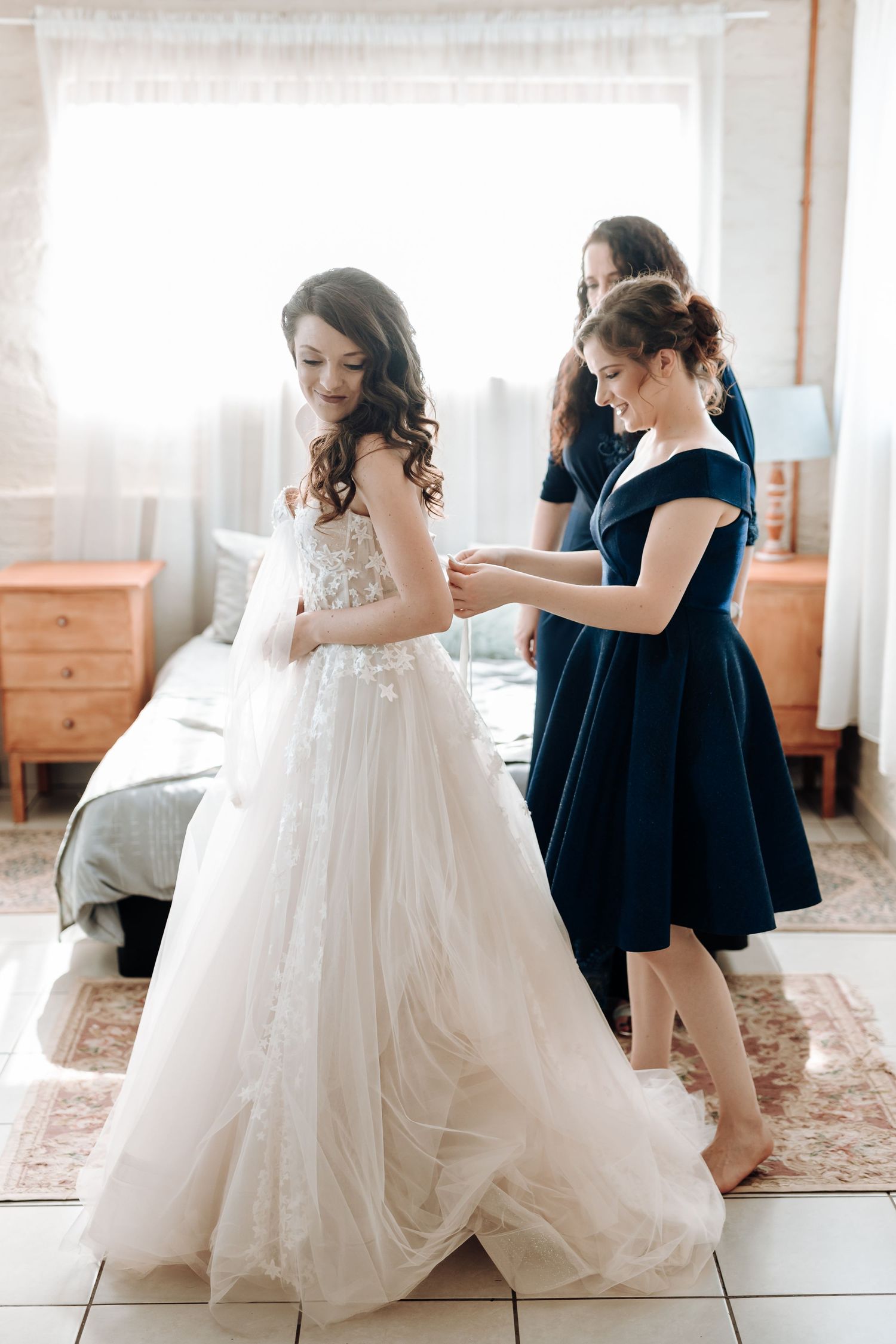 Women helping with final adjustments of a flowing white wedding dress in a sunlit room.