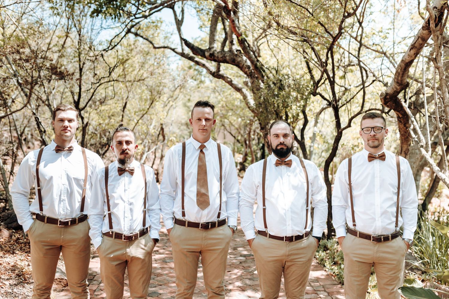 Groomsmen in matching tan pants and white shirts with suspenders pose for a formal photo.