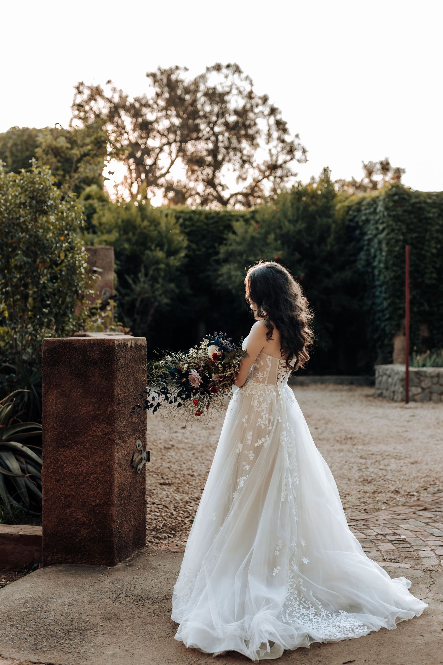 A white wedding dress with lace detail and train photographed from behind in a garden setting.