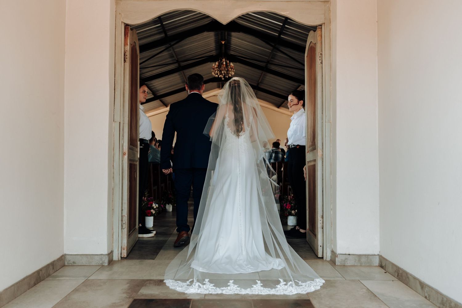 Looking down a white corridor, a couple stands at the entrance of a door during a wedding ceremony.