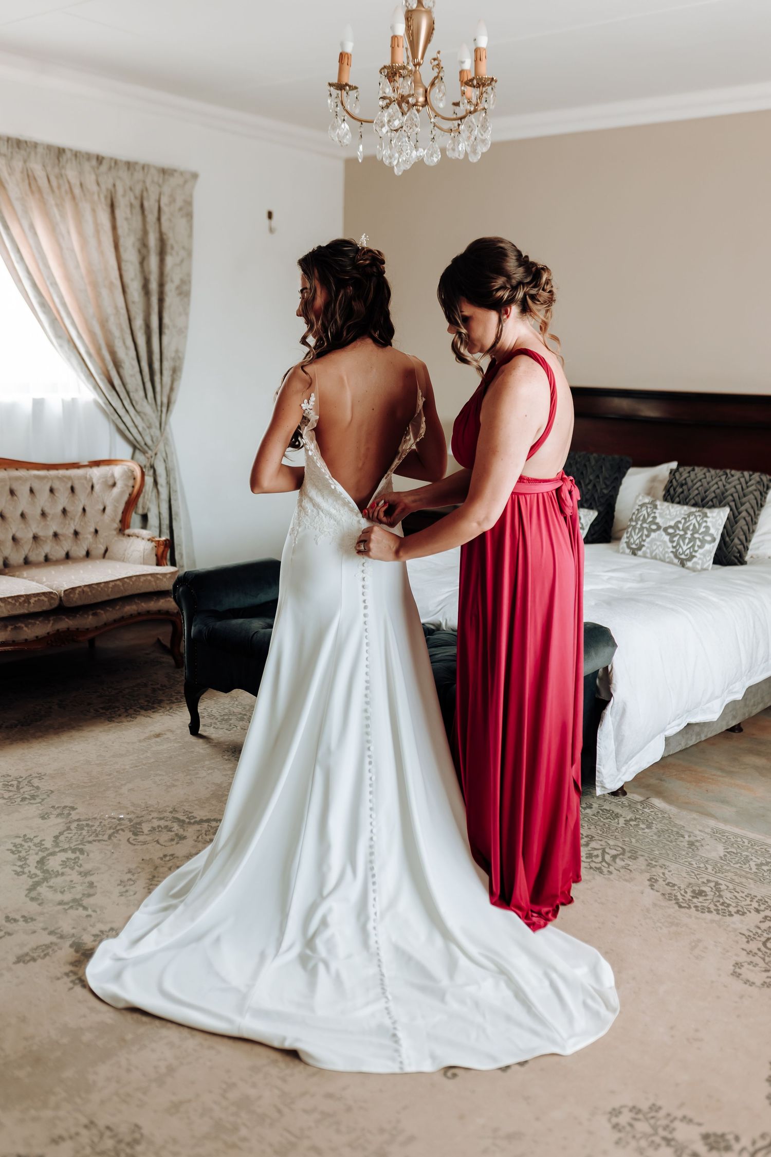 A wedding dress being buttoned up in an elegant bedroom with crystal chandelier and neutral decor.