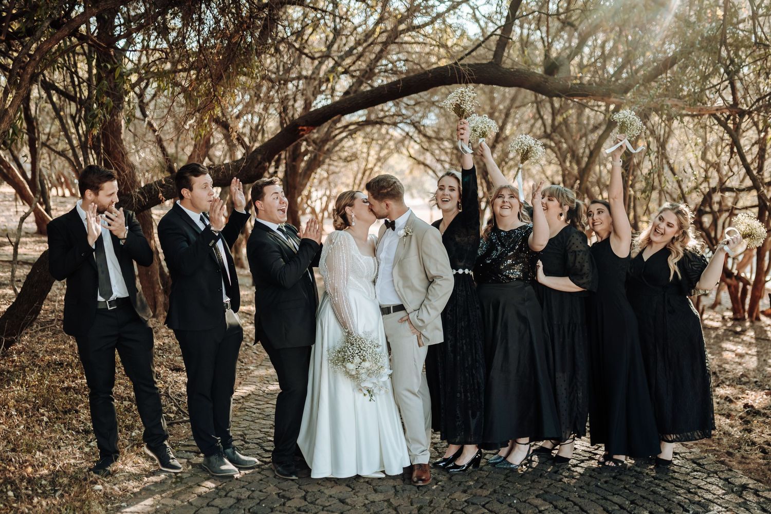 Wedding group celebrates joyfully beneath tree branches in a natural forest setting.