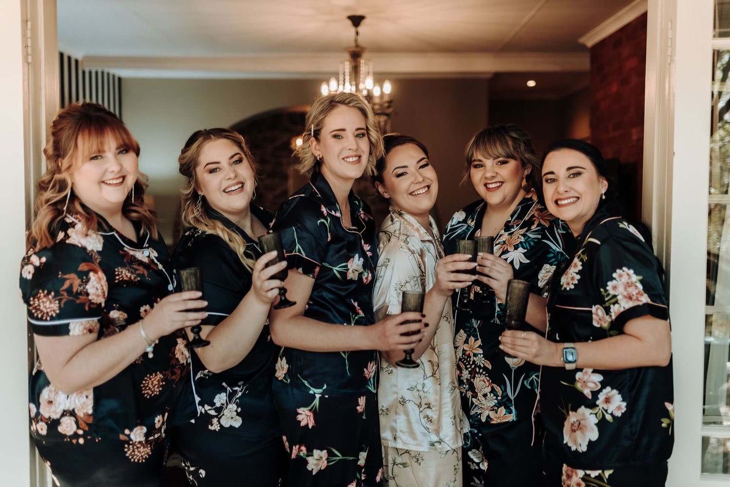 Bridal party wearing matching floral black robes holding drinks during wedding preparations.