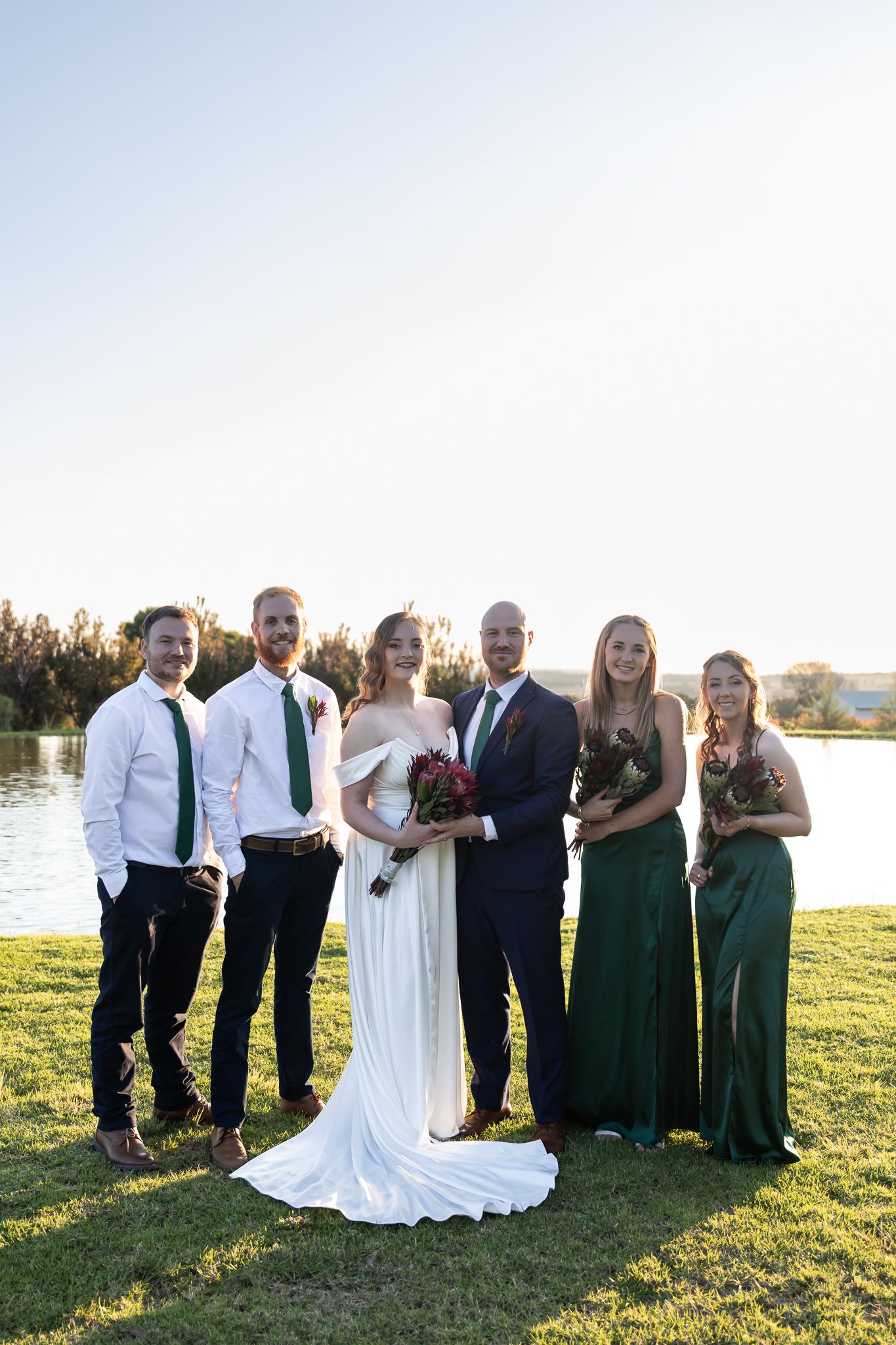 Wedding party posing together on grassy lawn with waterfront view at sunset.