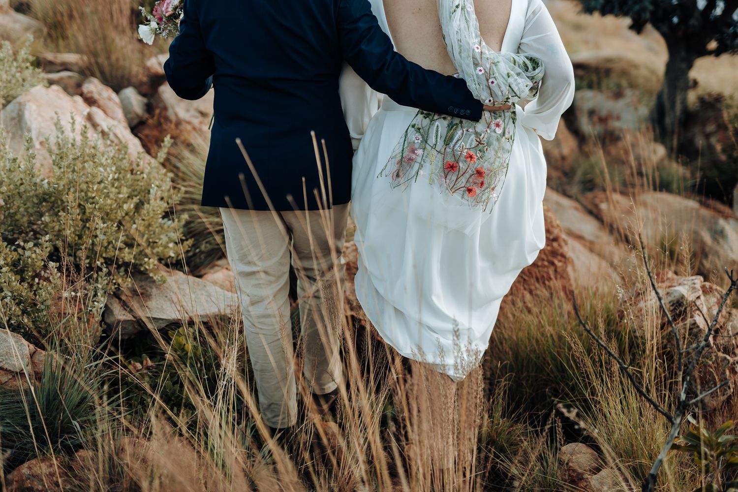A couple's lower bodies shown in formal attire standing among tall grass and rocks in a natural outdoor setting.