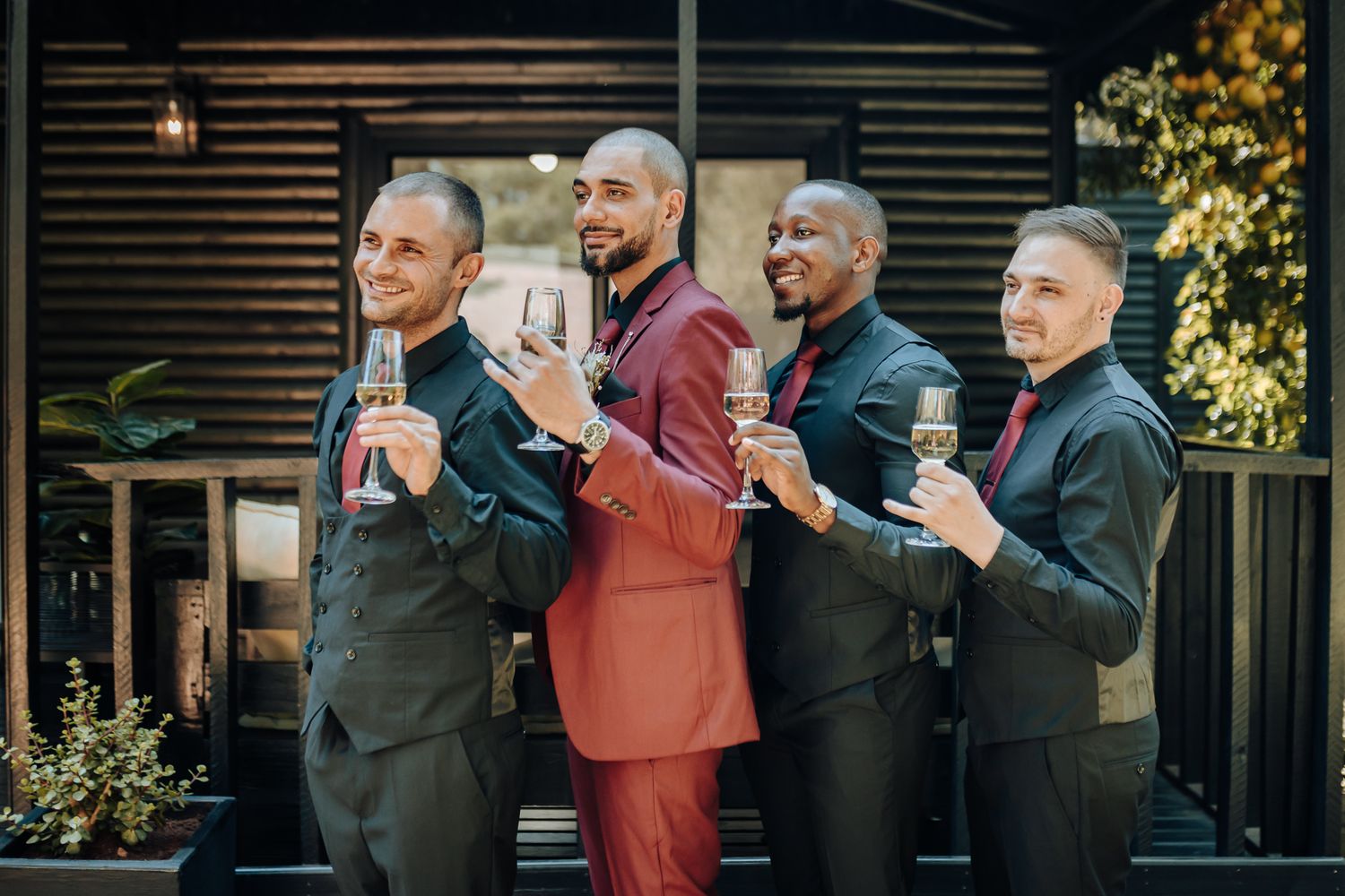 Groomsmen in formal suits posing together with drinks against a rustic wooden backdrop.