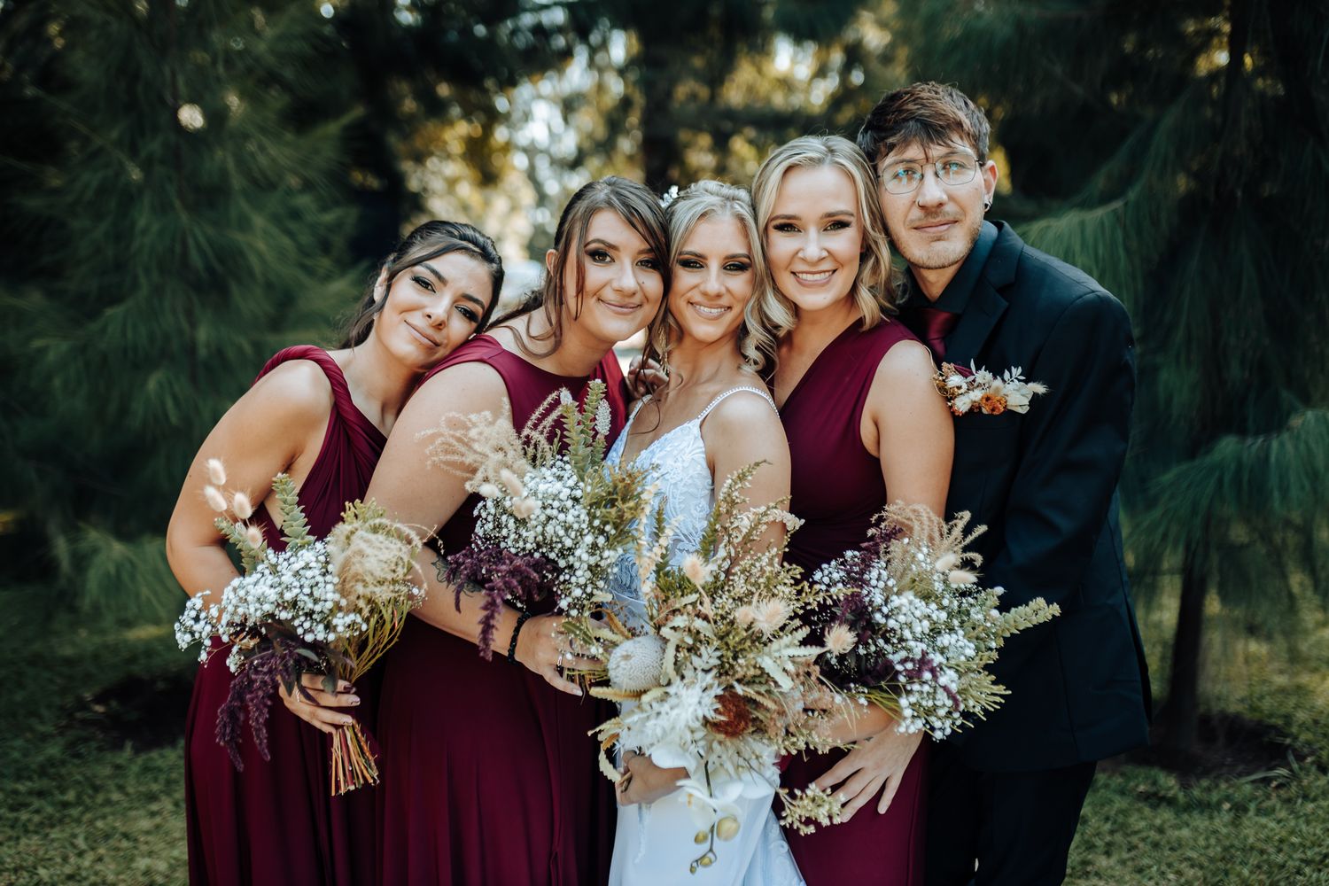 Wedding party in burgundy dresses and black suit pose together with dried flower bouquets.