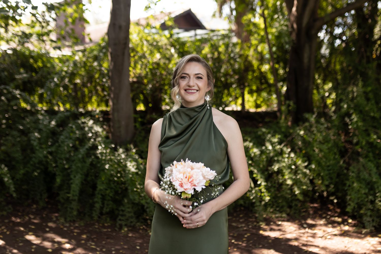 A person in a sage green dress holds a pink bouquet while standing in dappled sunlight.